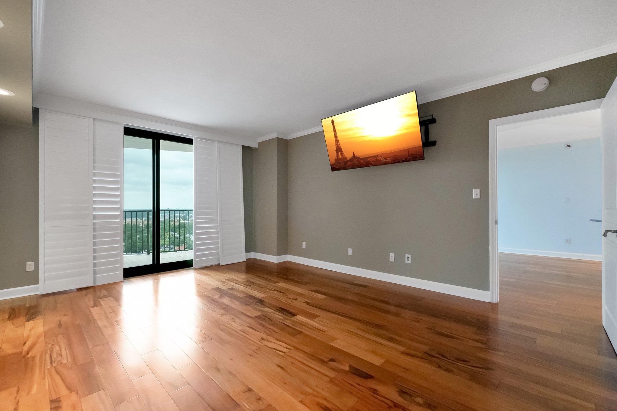 Empty living room with glossy wood floors, balcony doors, white shutters, and a wall-mounted television.