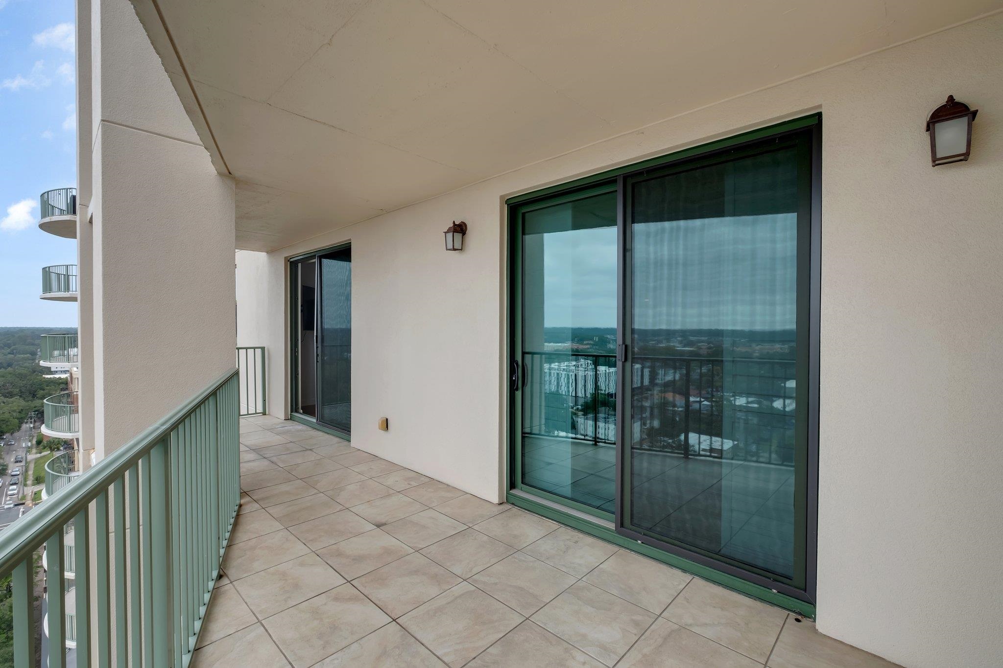 High-rise balcony with tiled flooring, green railings, and sliding glass doors overlooking the city.