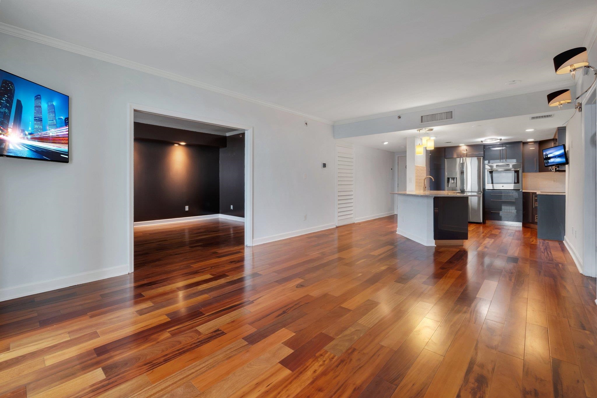 Open-plan condo living area and kitchen with multitone hardwood floors and dark cabinetry.