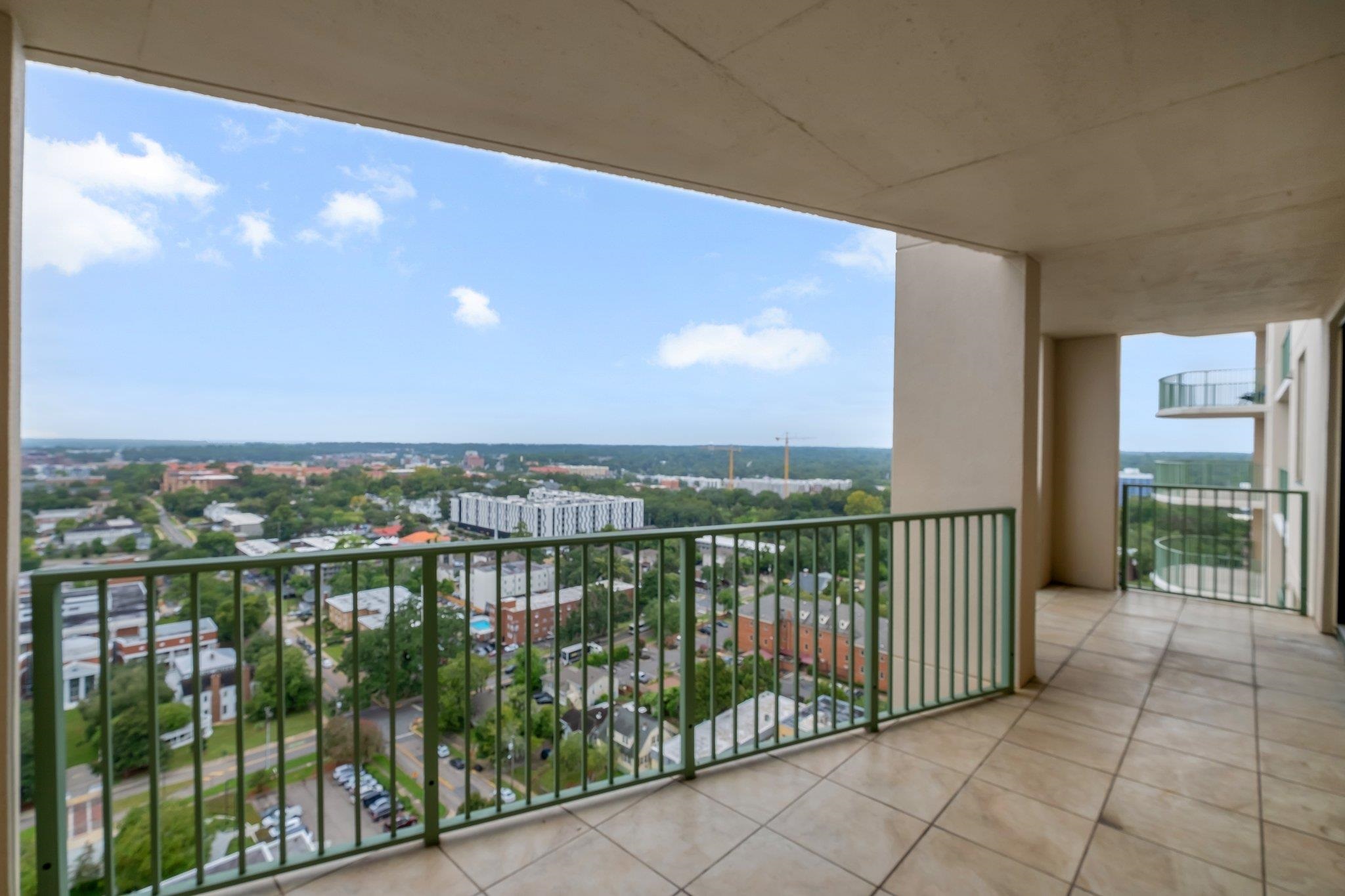 High-rise balcony overlooking a leafy neighborhood, with tiled flooring and dark green railings.