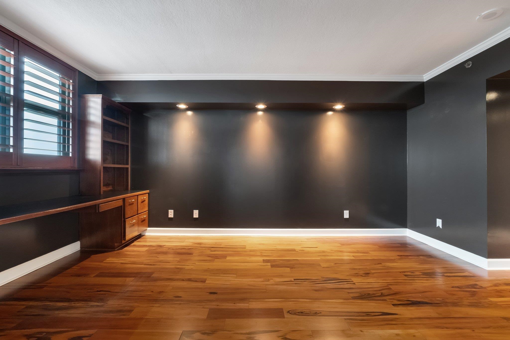 Empty room with a built-in wooden desk, shelving, dark walls, and hardwood floors.