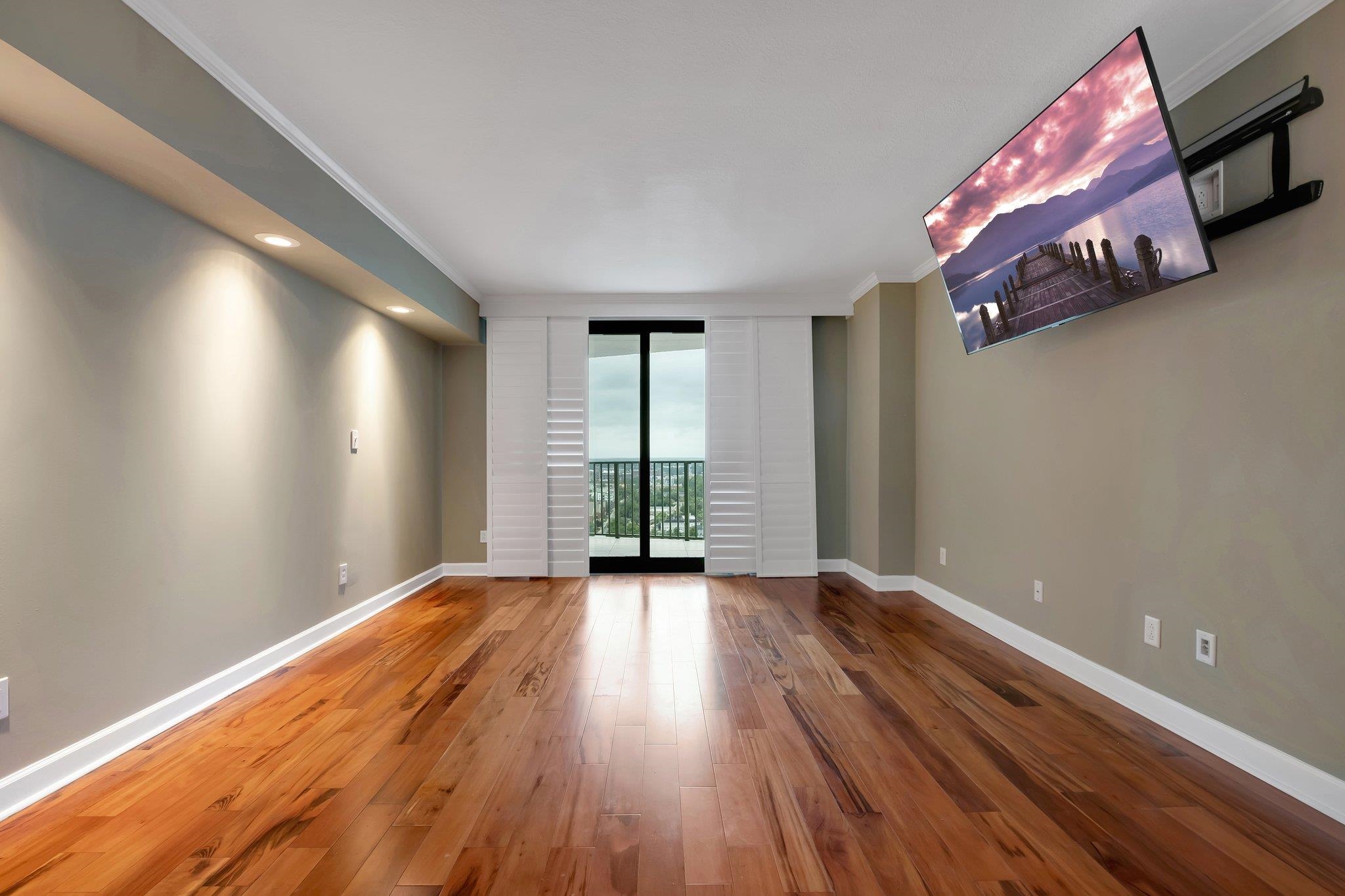 Unfurnished condo living room with hardwood floors, white shutters, balcony doors, and a wall-mounted television.