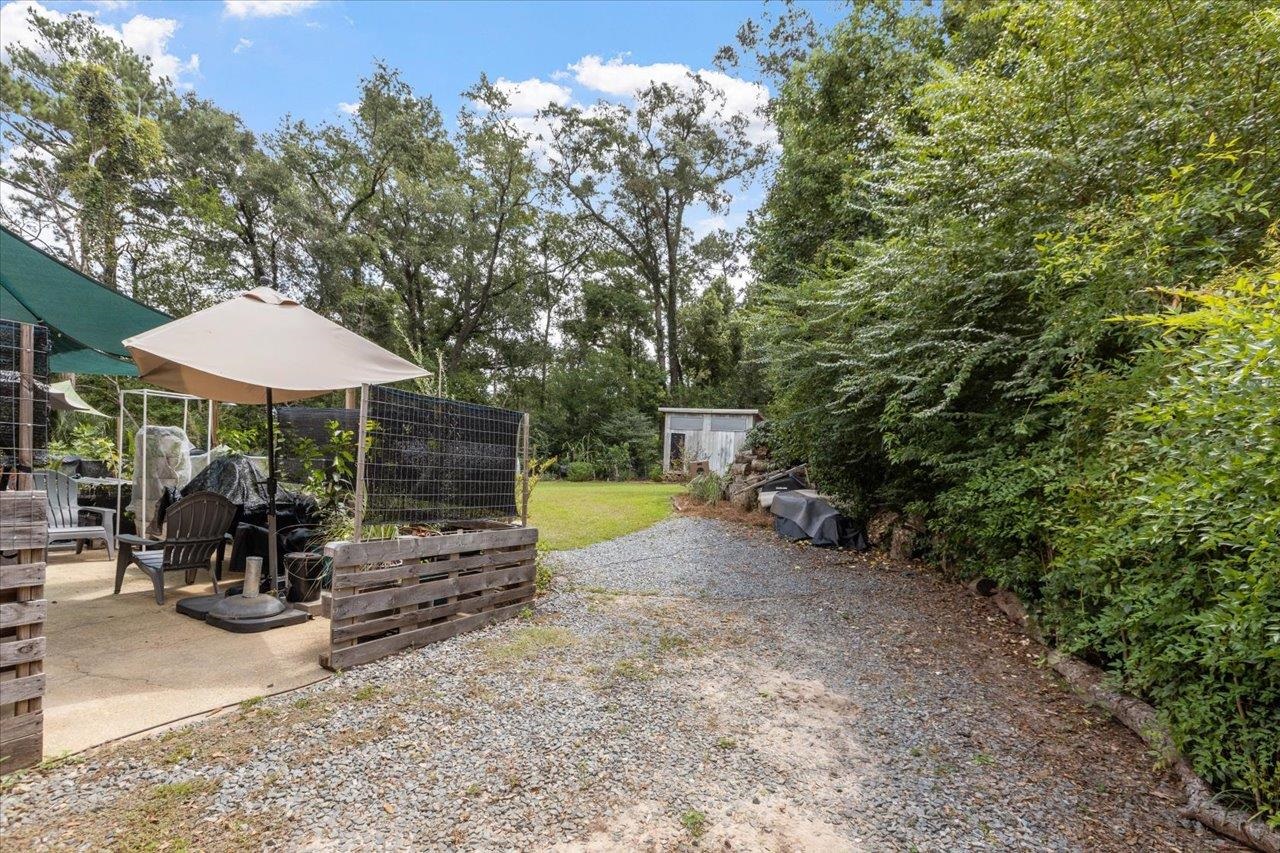 Gravel driveway beside a shaded patio with umbrella seating and a small backyard shed.