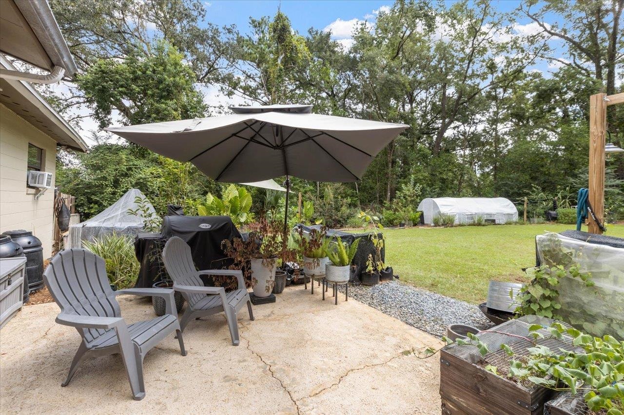 Backyard patio with gray chairs under a large umbrella, potted plants, raised beds, and greenhouses.