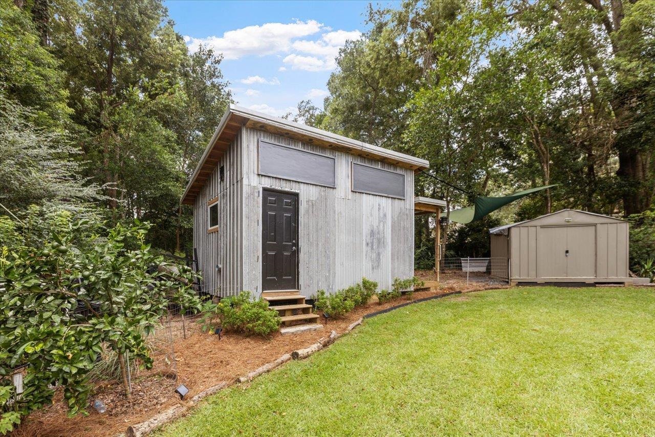 Weathered wood outbuilding with a dark door and high windows beside a storage shed in a leafy backyard.