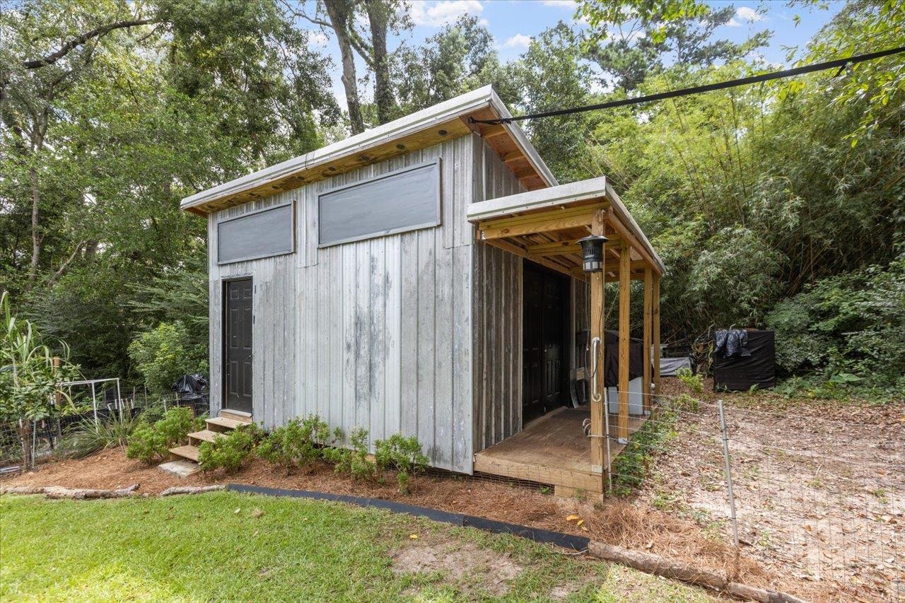 Small gray wood-sided outbuilding with high windows and a covered porch among trees.