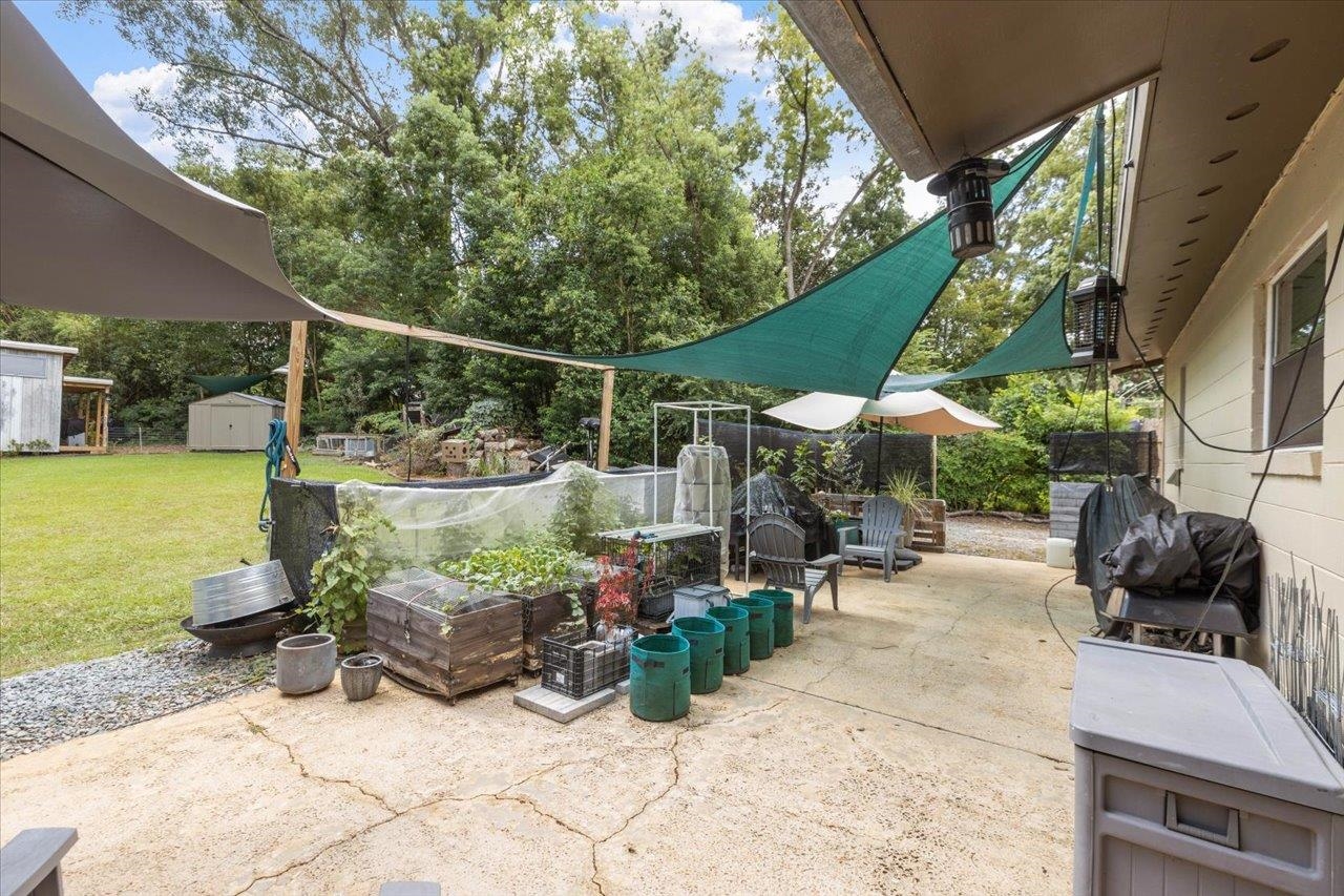 Back patio with shade sails, raised garden beds, outdoor chairs, and a tree-lined lawn.