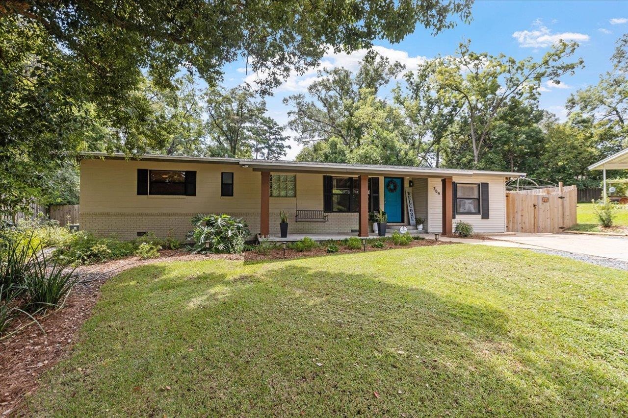 Single-story home with a blue front door, covered porch, and broad lawn beneath mature trees.