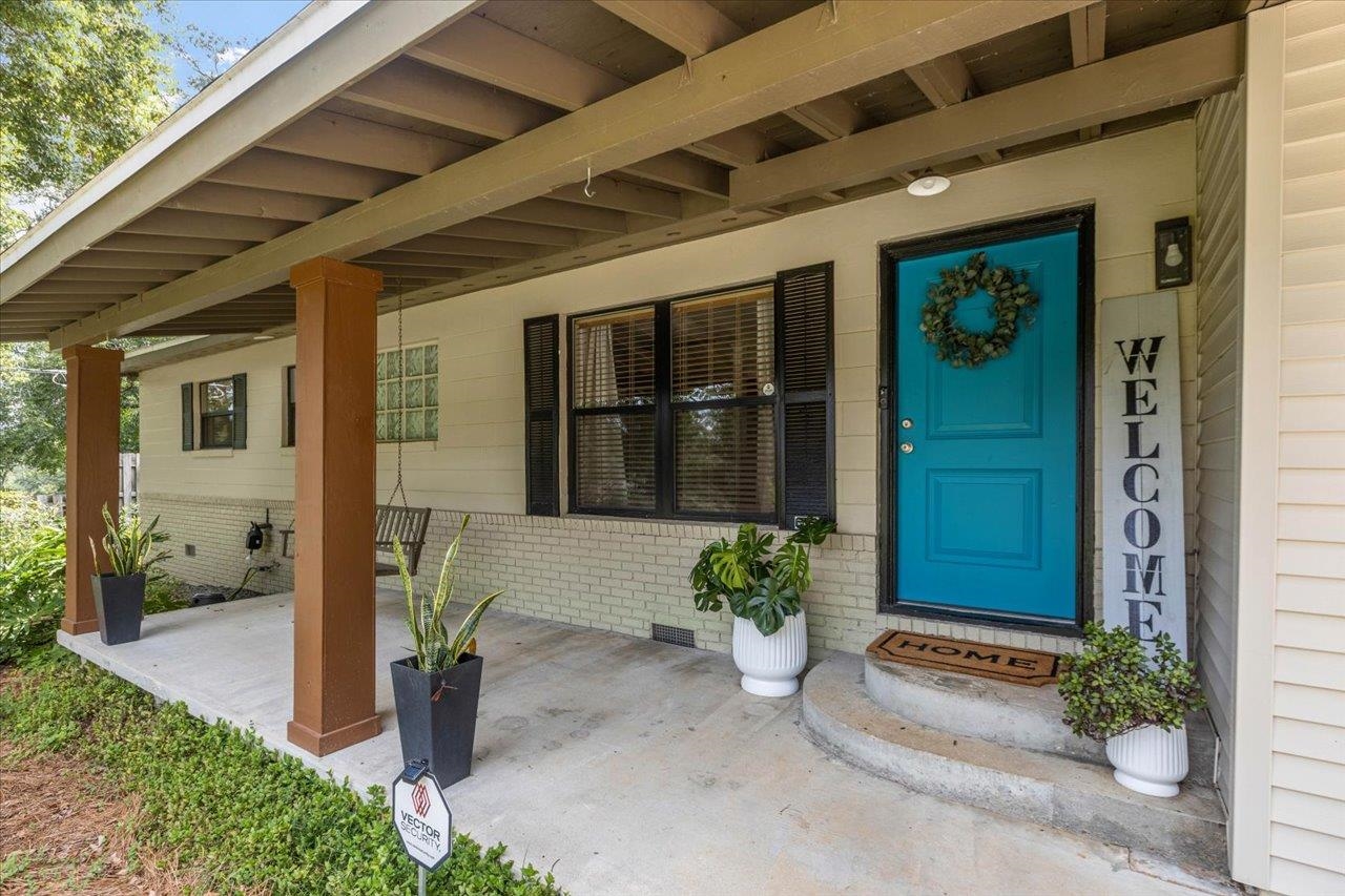 Covered front porch with a bright blue door, wreath, potted plants, and welcome sign.