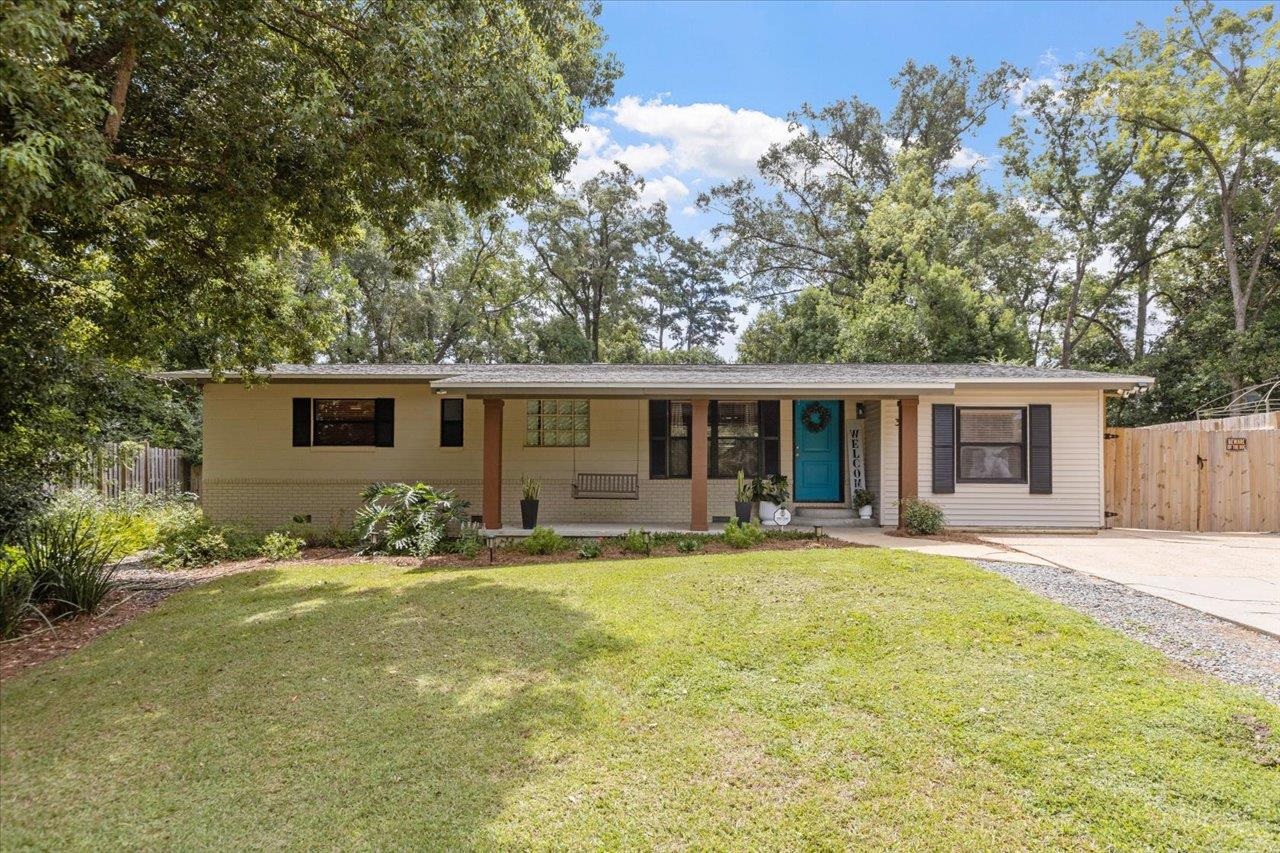 Single-story ranch house with a covered porch, teal front door, and lawn beneath mature trees.
