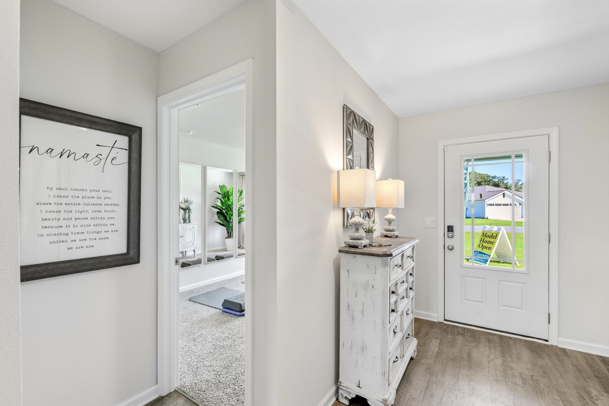 Staged entry foyer with a distressed white console, paired lamps, framed “namasté” print, and windowed front door.