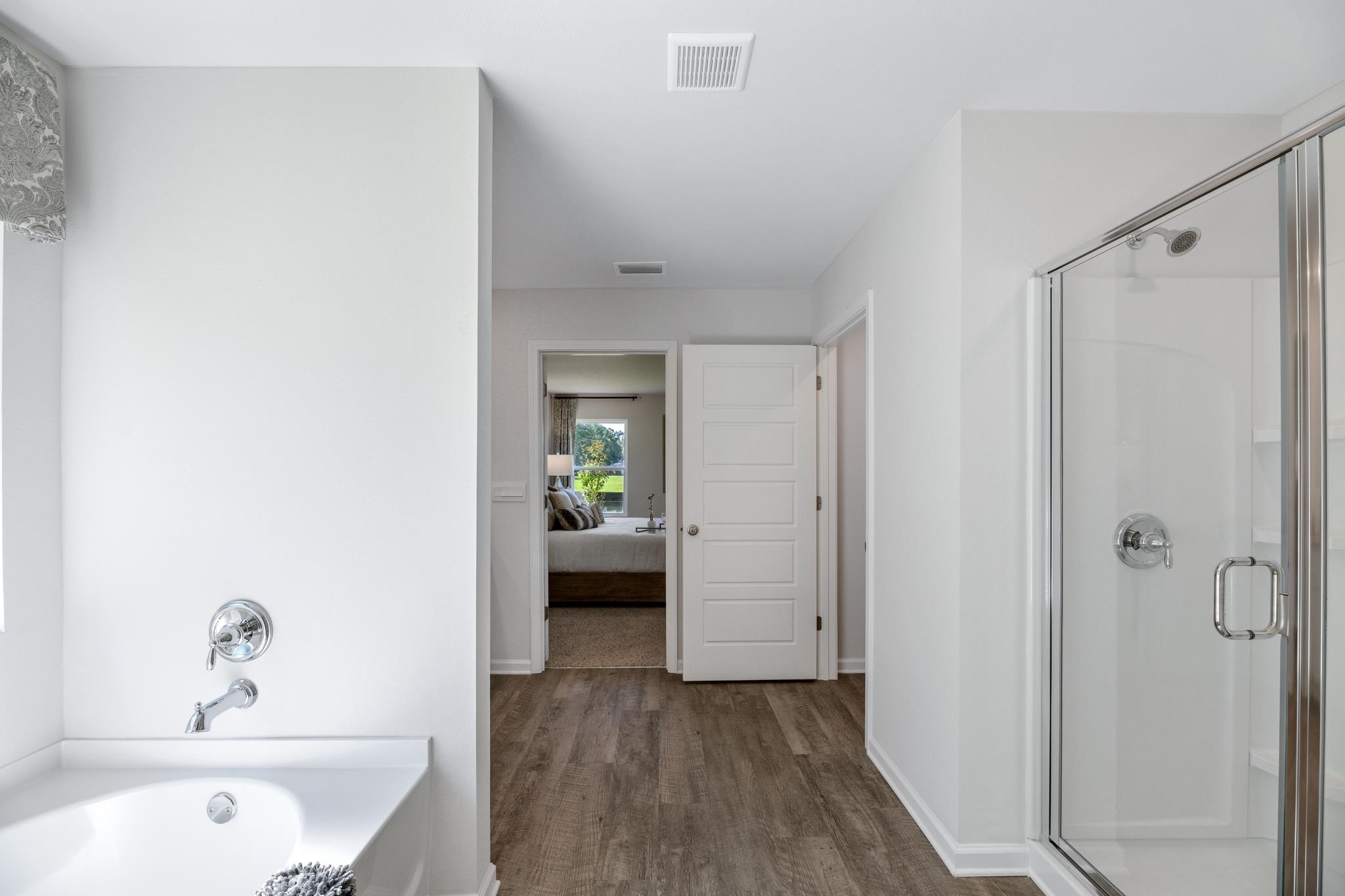 Bathroom with a soaking tub, glass shower, and wood-look flooring leading to a bedroom.