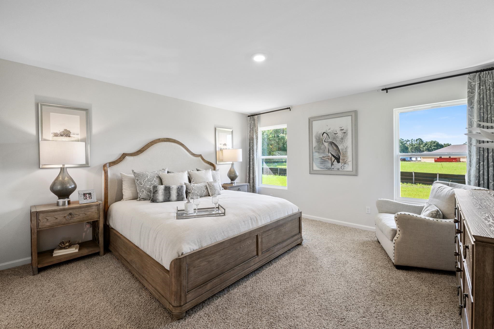 Staged bedroom with a large wood-framed bed, neutral bedding, and windows overlooking a lawn.