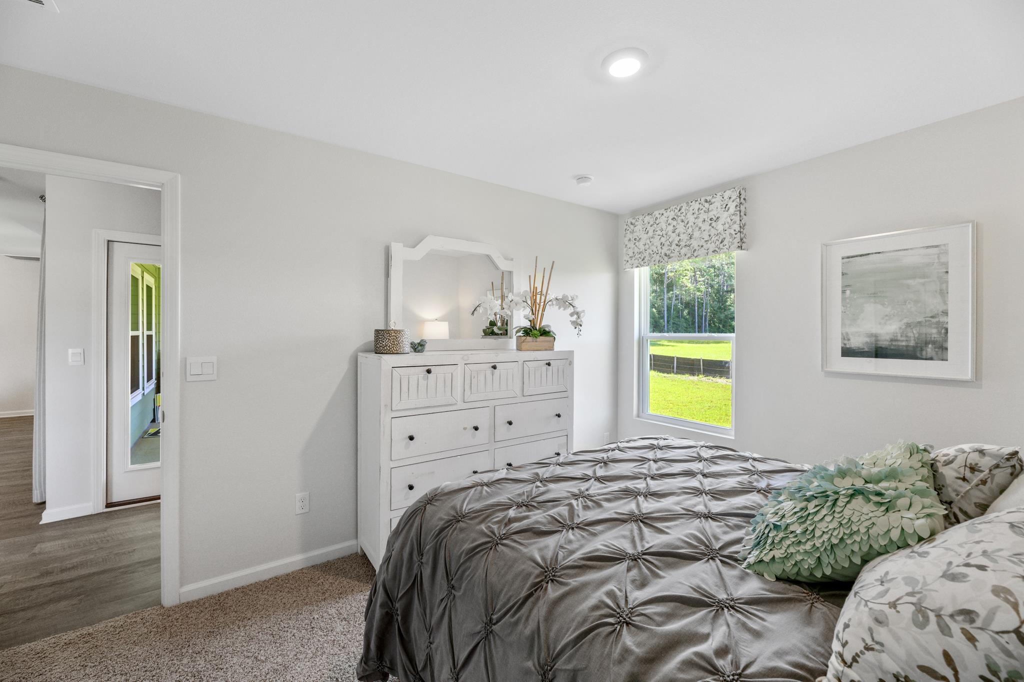Staged bedroom with a gray tufted bed, white dresser, and window overlooking a green lawn.