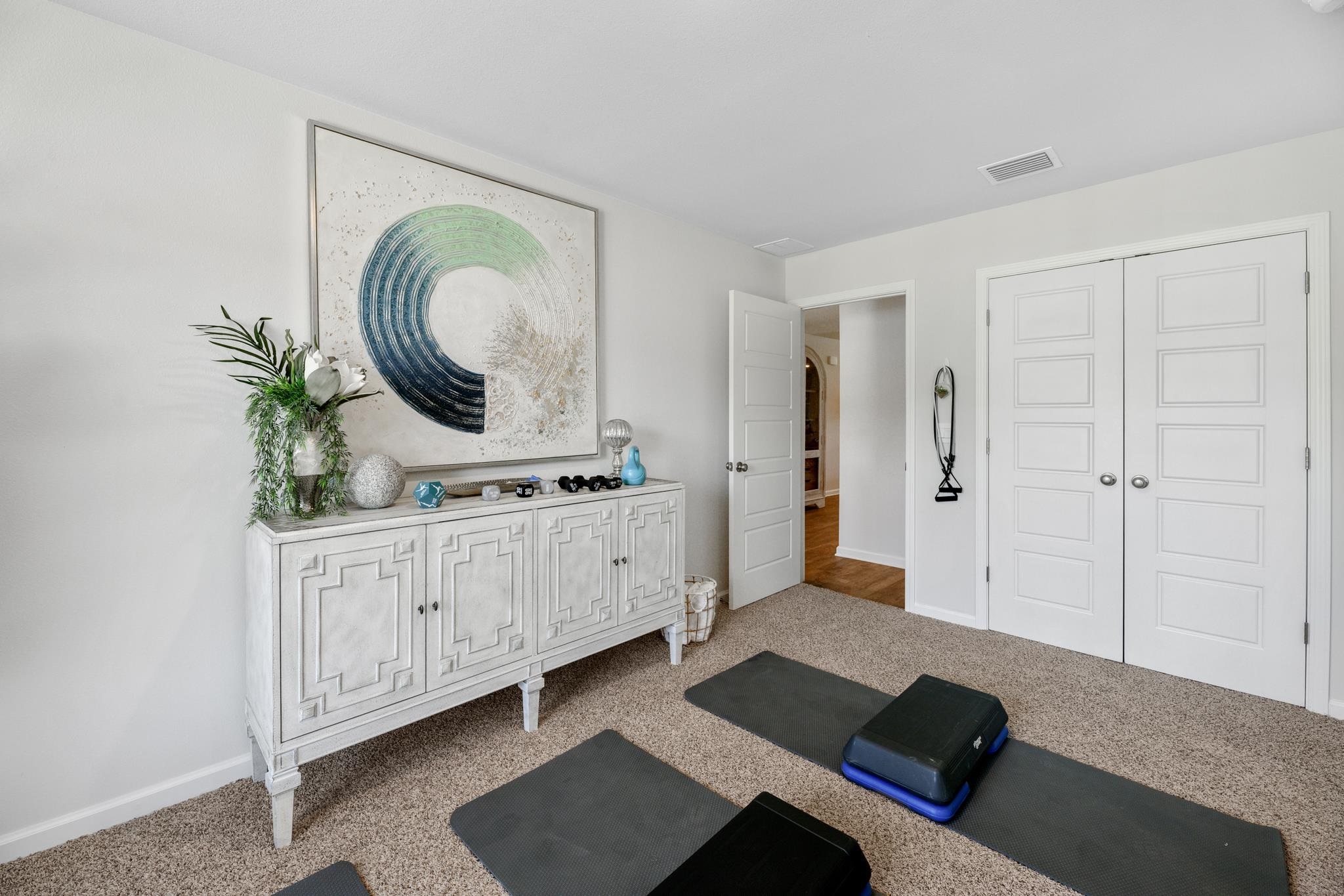 Carpeted home gym with exercise mats and step platforms beside a white cabinet and large abstract artwork.