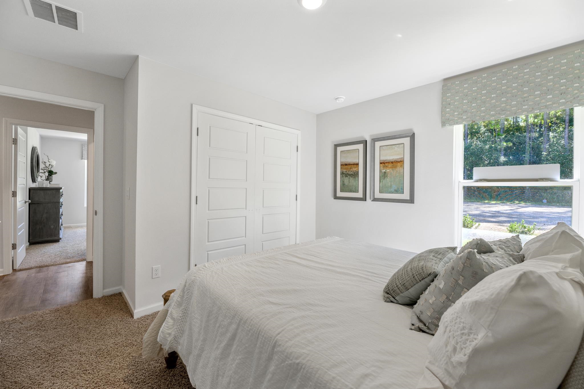 Furnished bedroom with a large white bed, paneled closet doors, and a window overlooking trees.