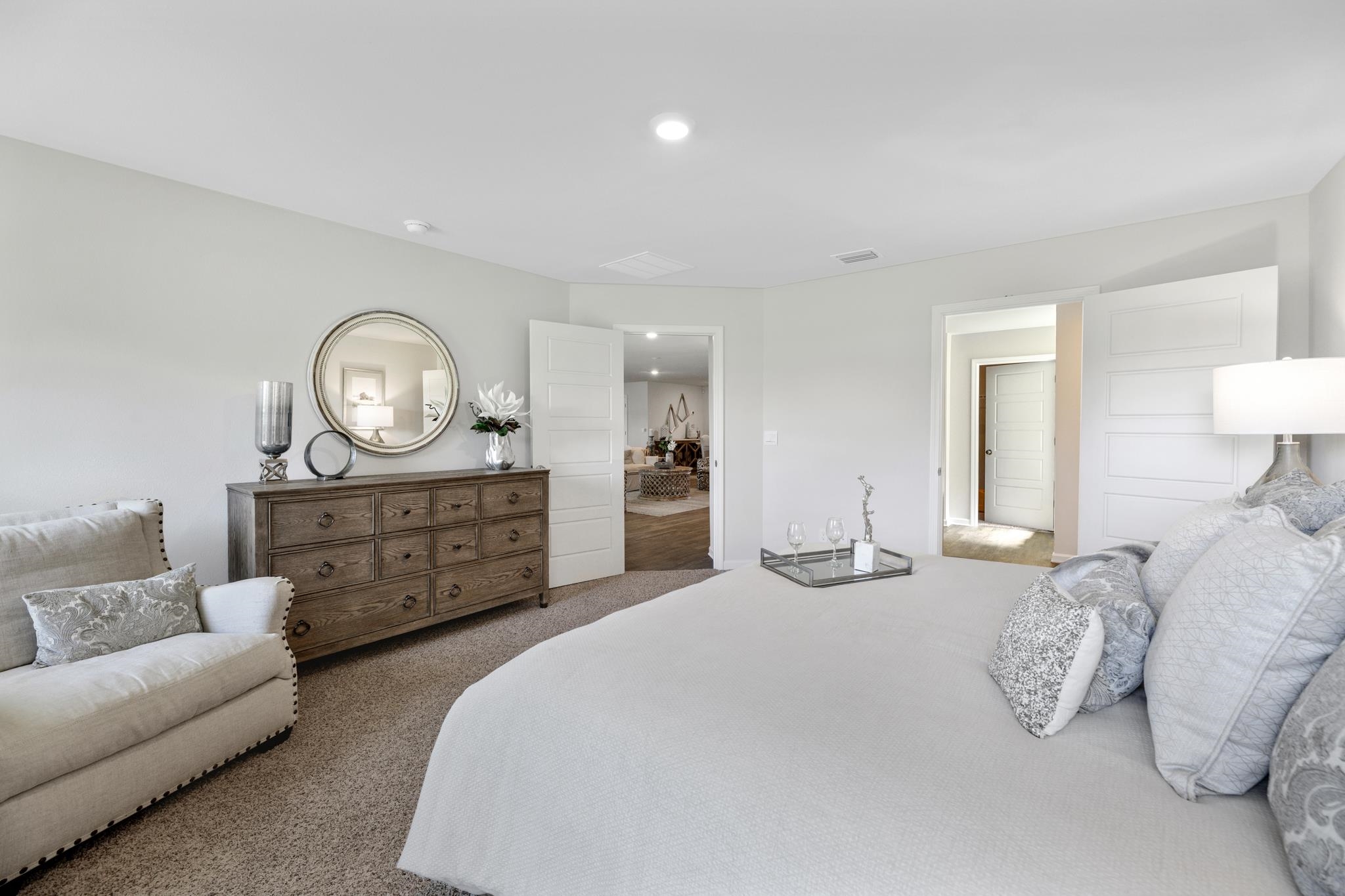 Bedroom with a large white bed, patterned pillows, and a dark wood dresser beneath a round mirror.