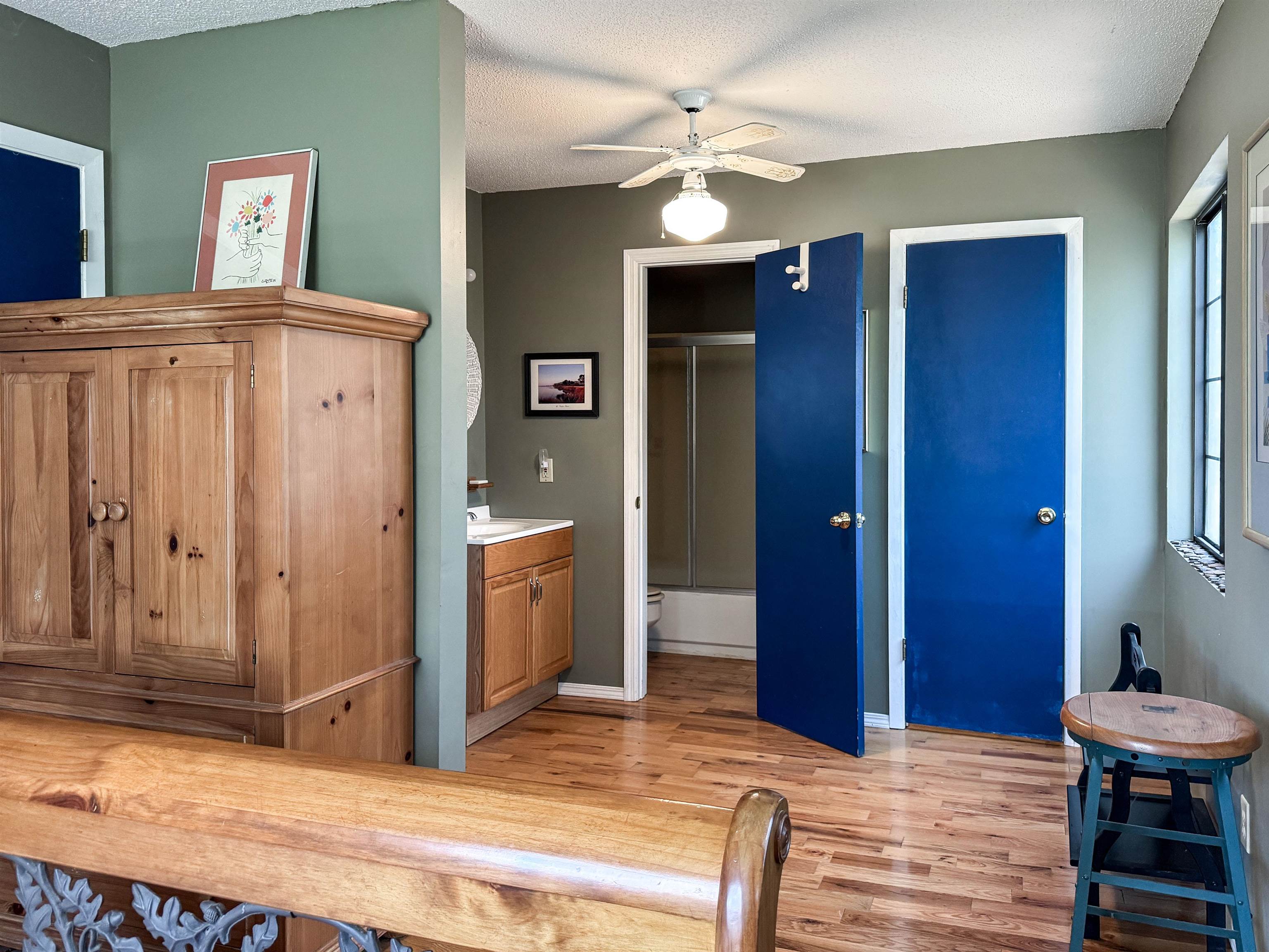 Bedroom with a large wooden armoire, hardwood floors, and bright blue doors beside an open bathroom.