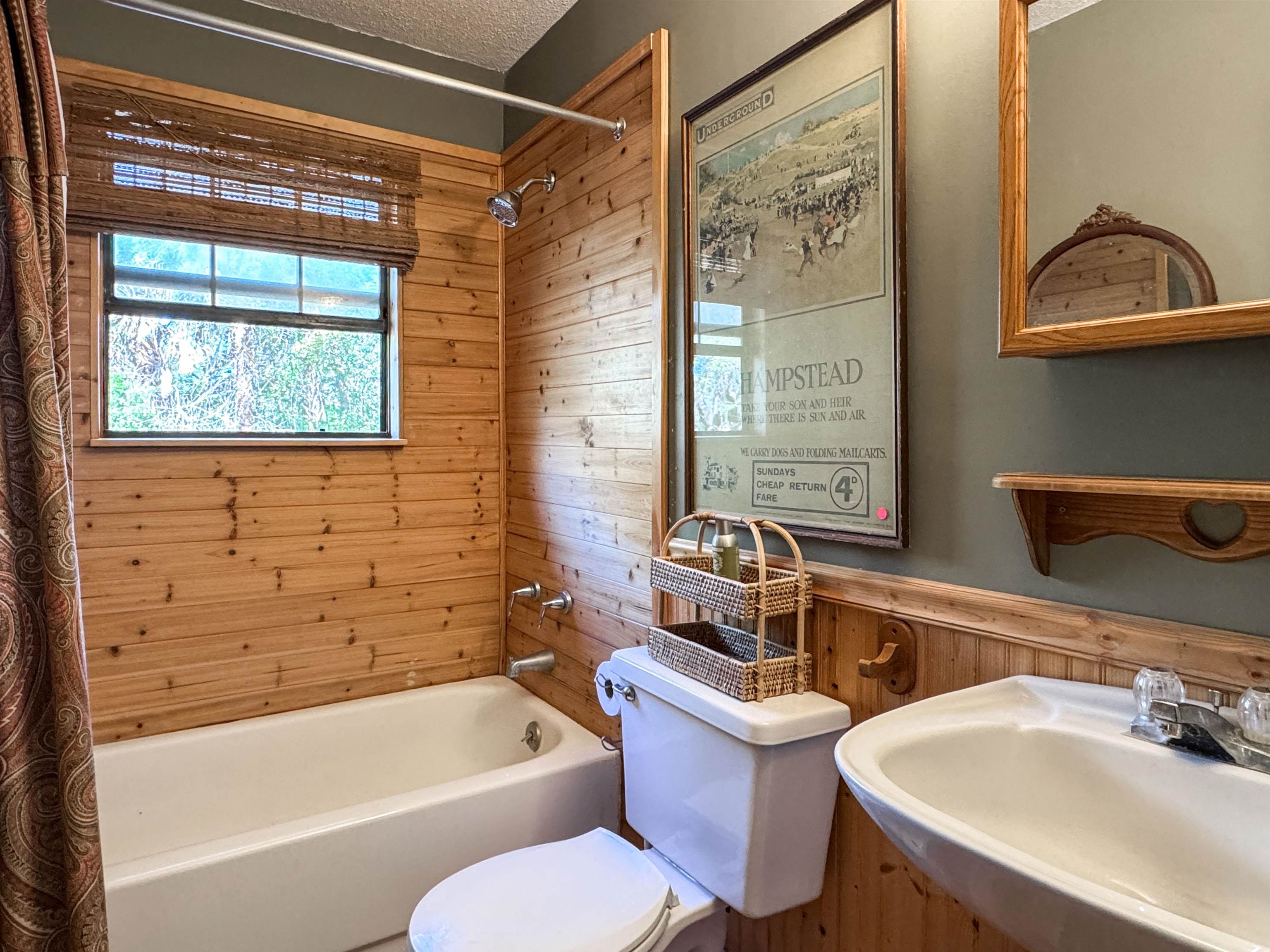 Bathroom with a white tub and pedestal sink, warm wood-paneled walls, and a window above the tub.