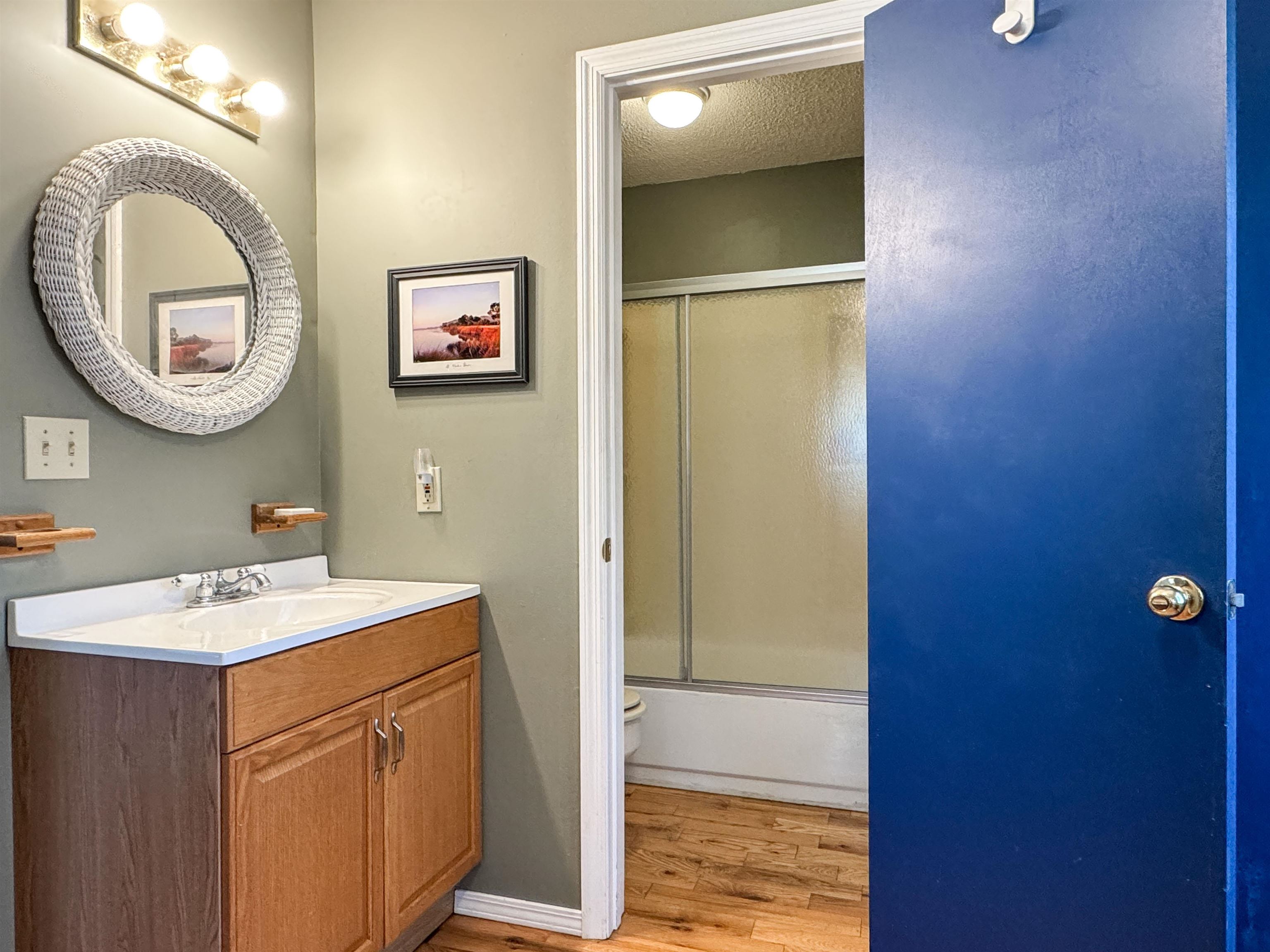 Bathroom vanity with a round woven mirror and a walk-in shower beyond the blue door.