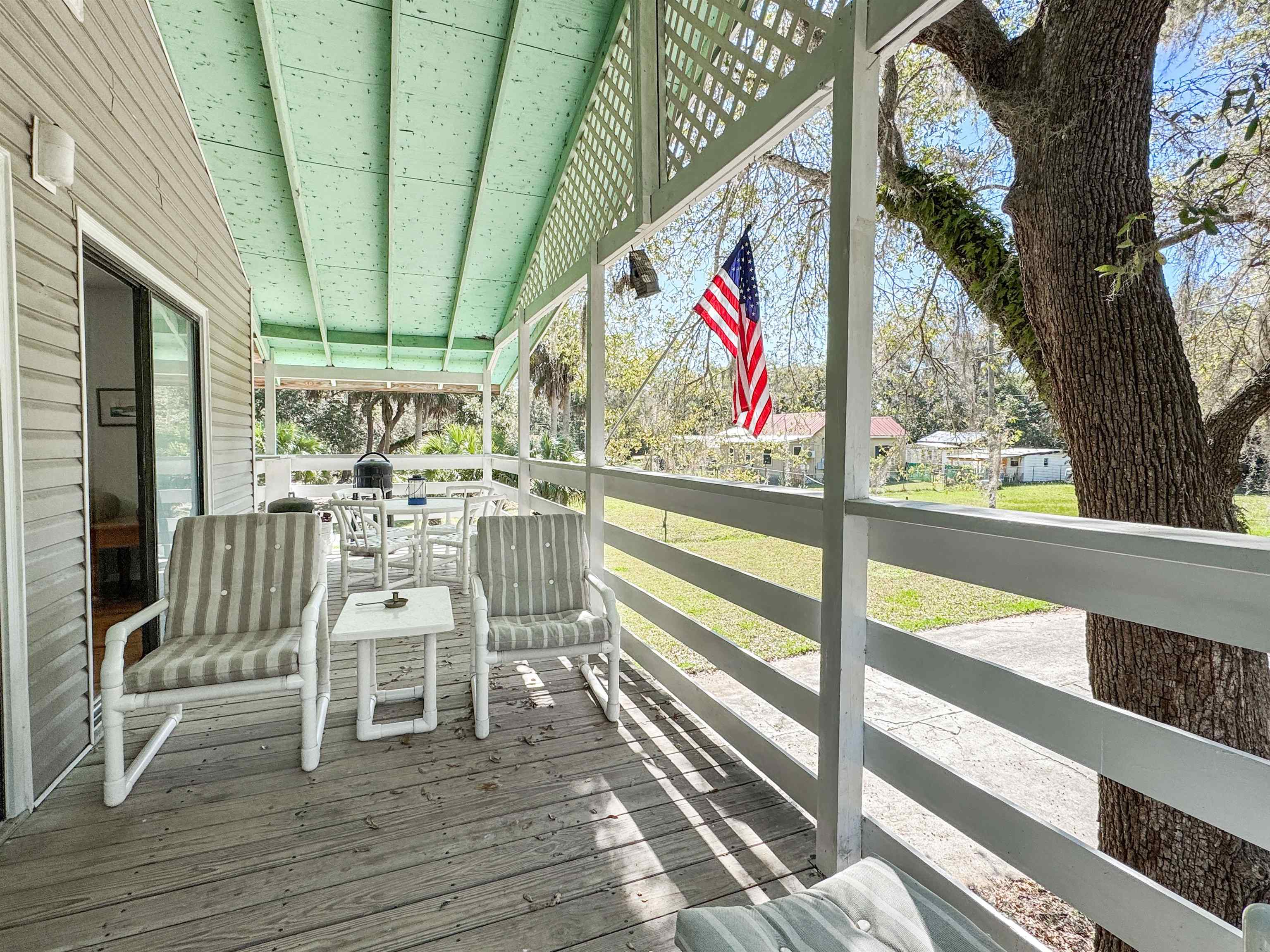Covered porch with striped chairs, an American flag, and a large oak tree beside the yard.