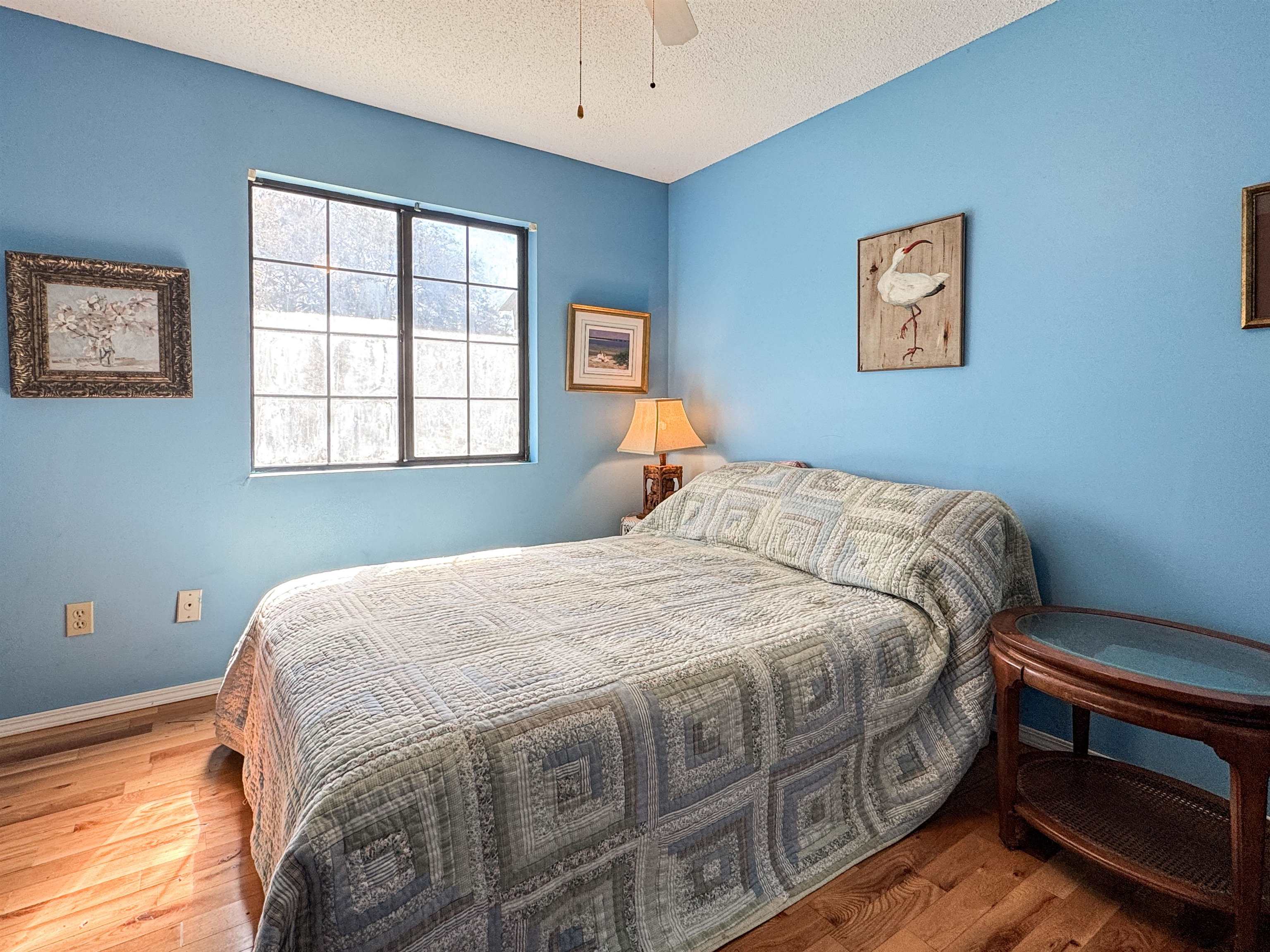 Bedroom with blue walls, a gray patchwork bedspread, hardwood floors, and a sunlit window.