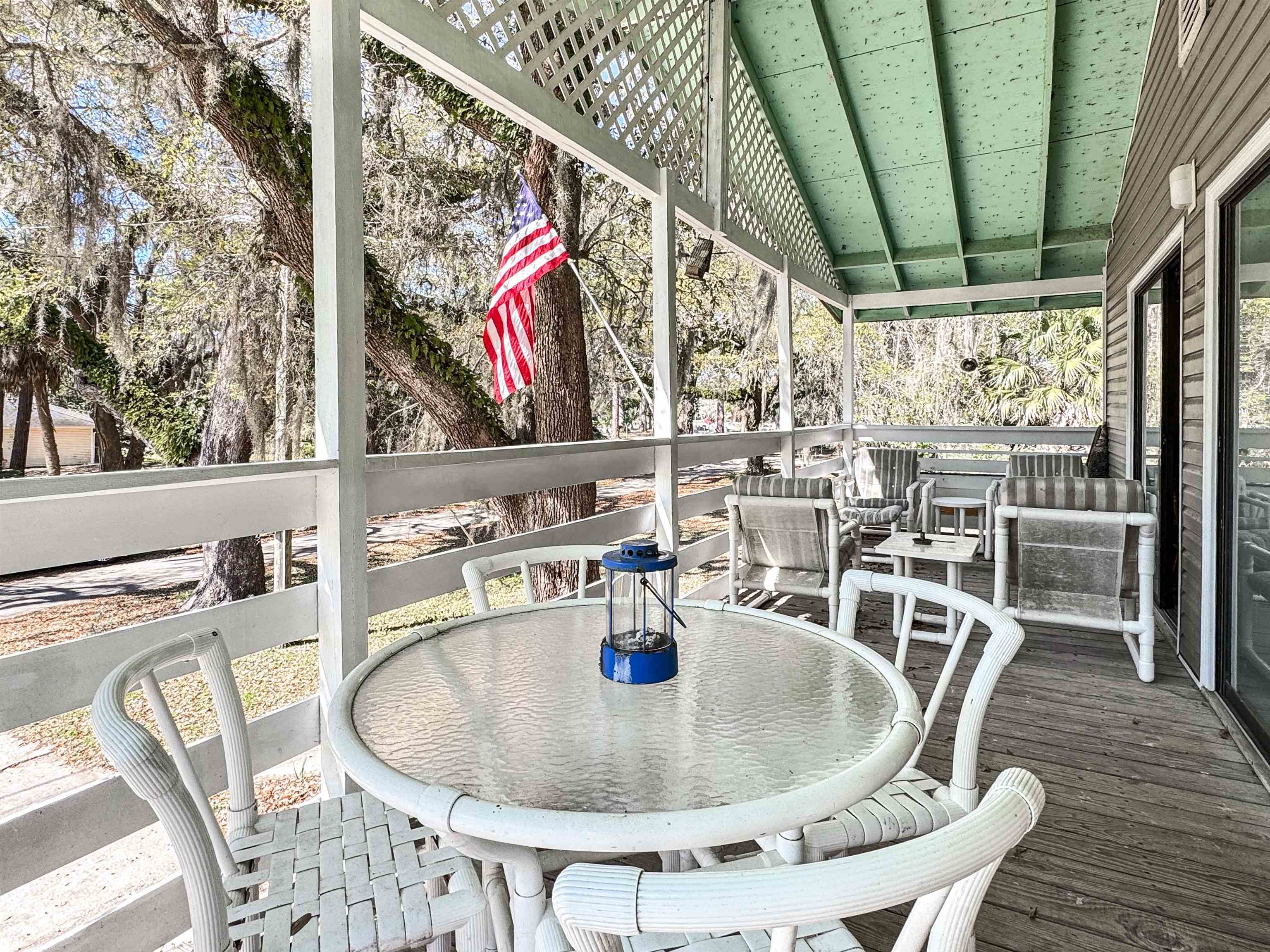 Covered porch with white wicker seating, a round table, and an American flag among mature trees.