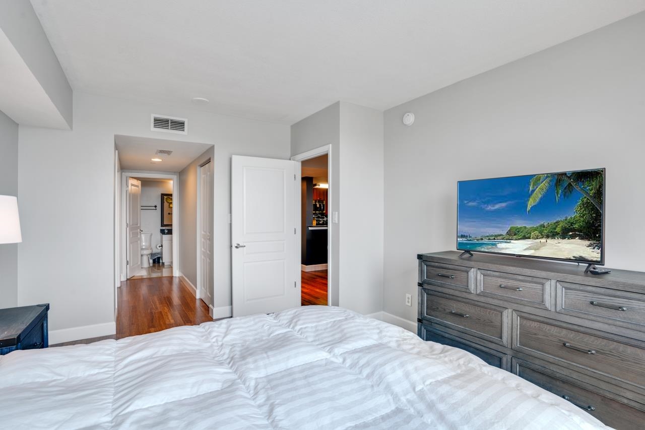 Bedroom with a white bed, gray dresser, and television showing a tropical beach.