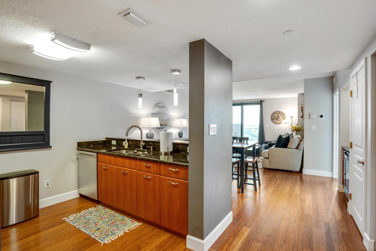 Condo kitchen with warm wood cabinets and dark countertops opening into a furnished living area.