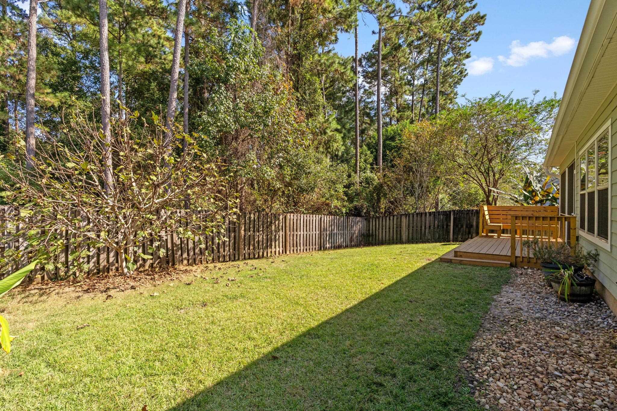 Fenced backyard with a sunny lawn, mature trees, and a small wooden deck beside the house.