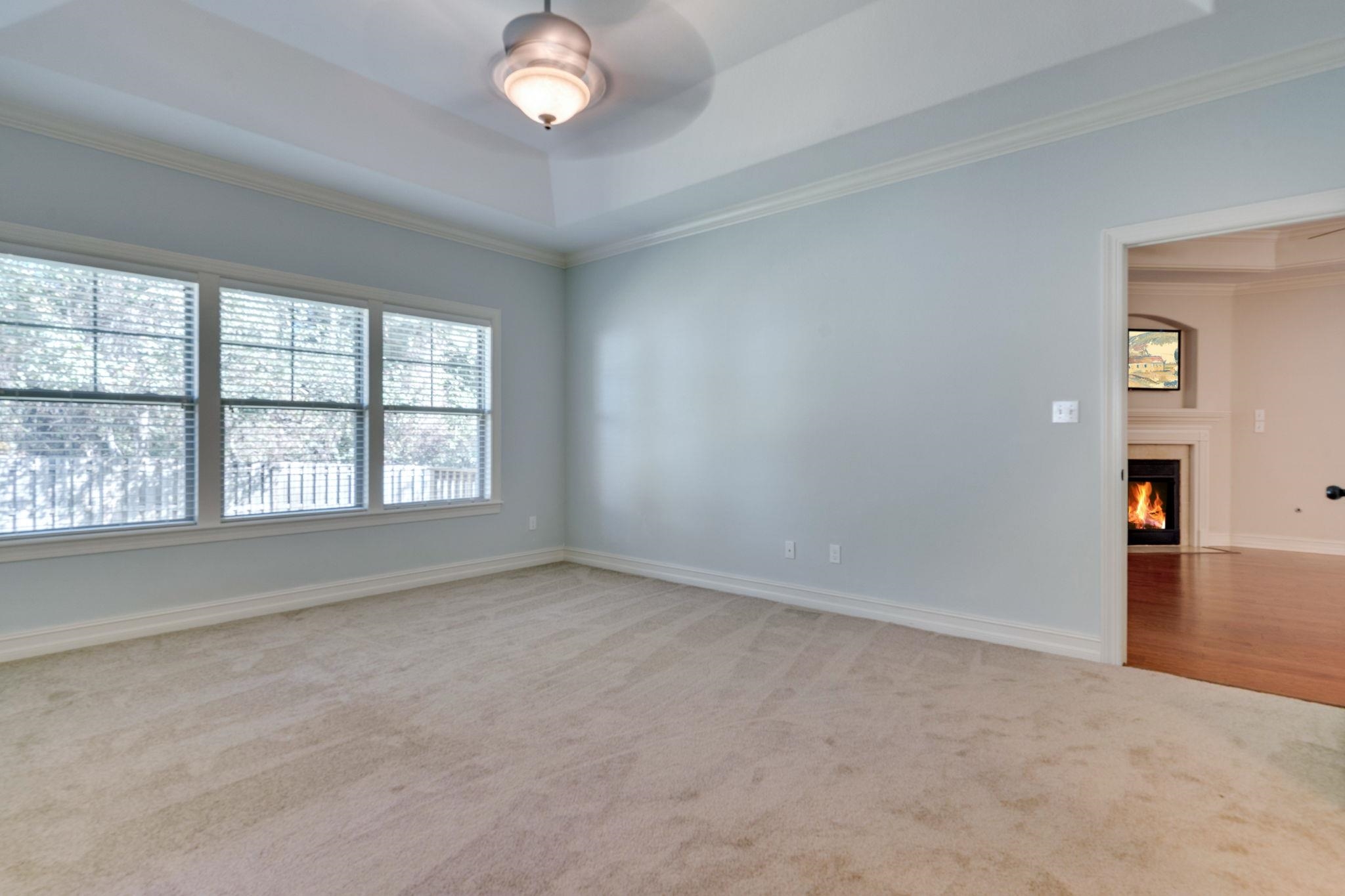 Empty carpeted room with three shaded windows and a doorway to a room with a lit fireplace.