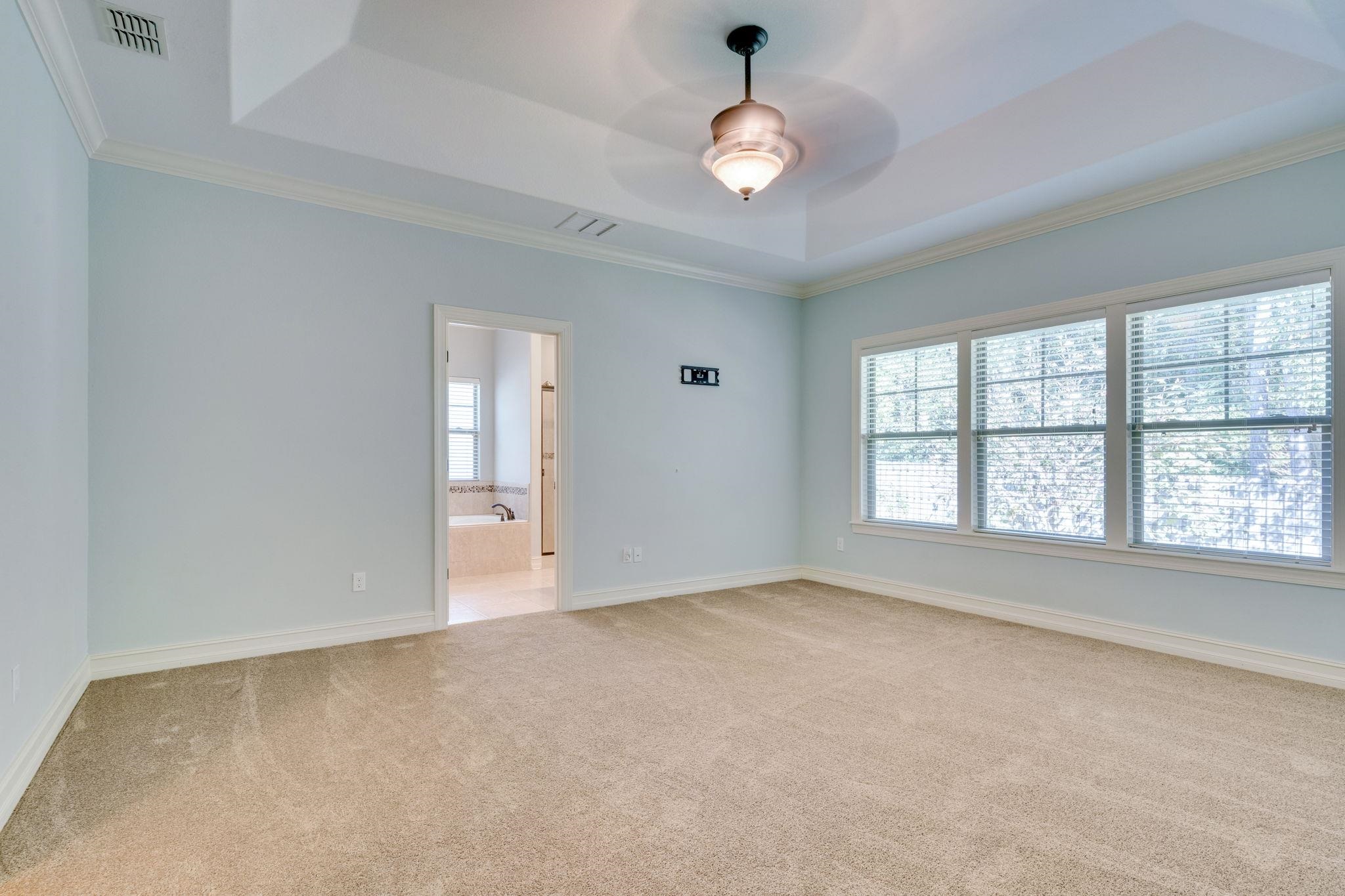Empty bedroom with a tray ceiling, ceiling fan, three wide windows, and an adjoining bathroom doorway.