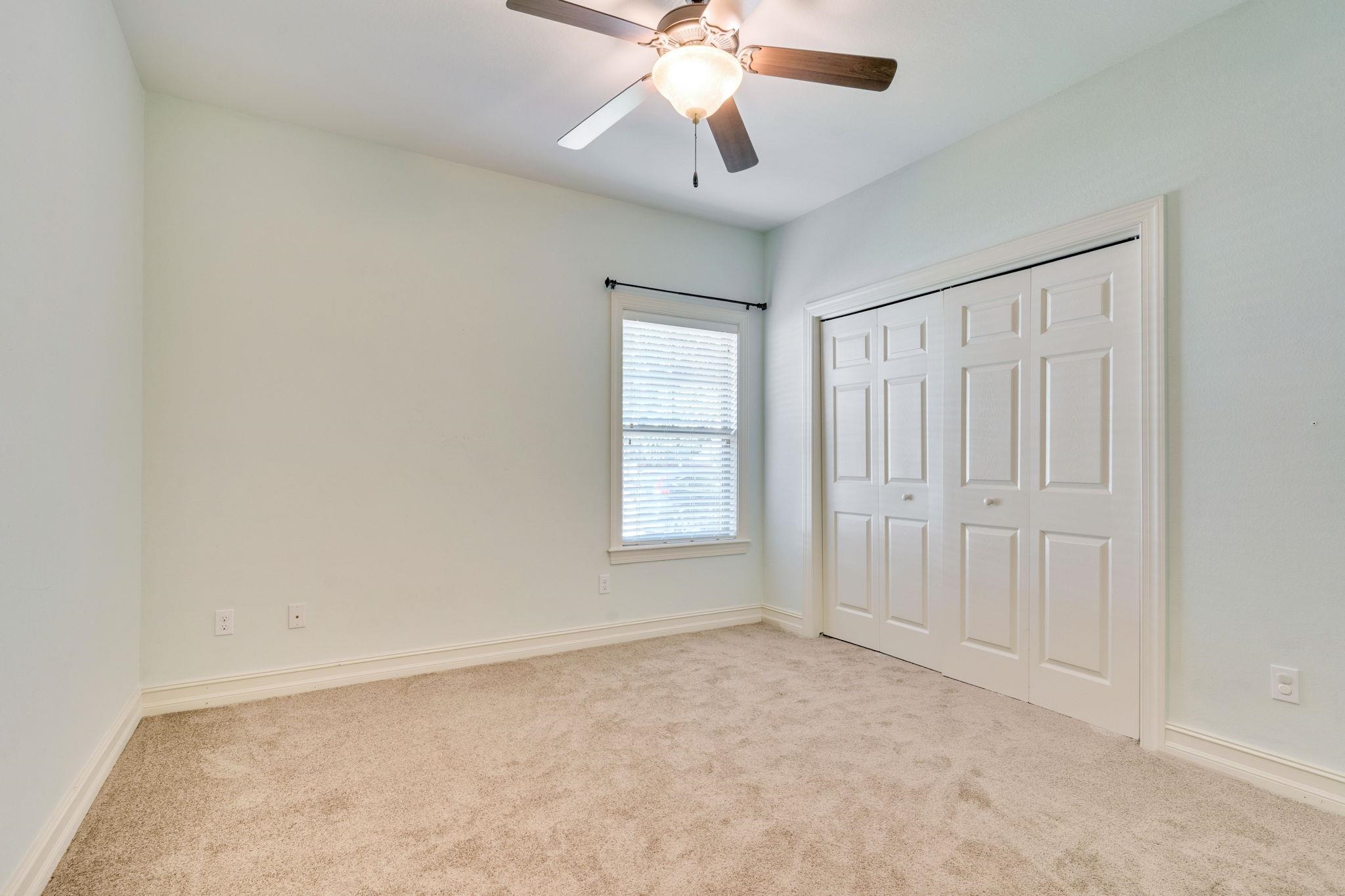 Empty bedroom with beige carpet, a window with blinds, and white paneled closet doors.