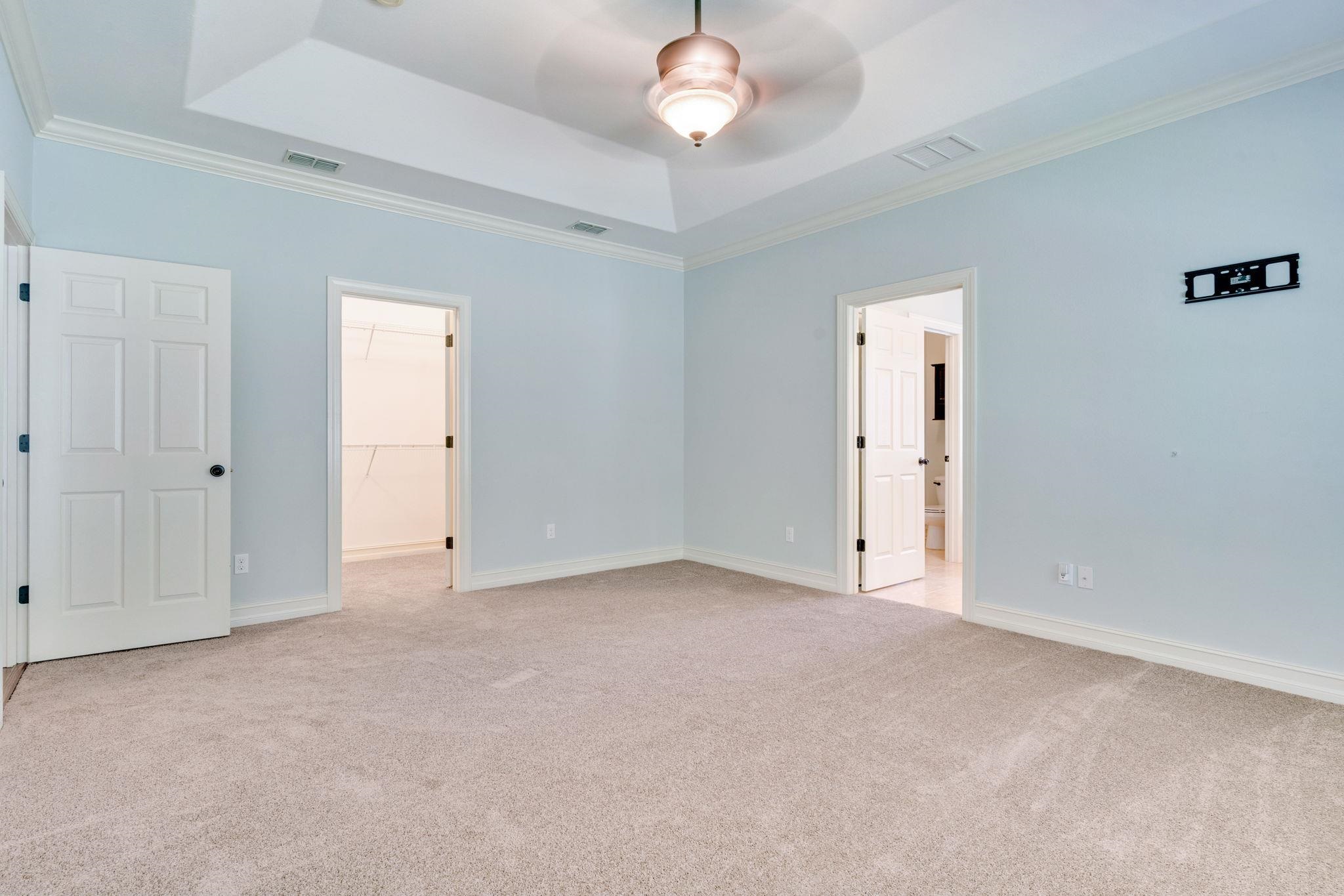 Empty bedroom with pale blue walls, beige carpet, a tray ceiling, and open doorways to a closet and bathroom.
