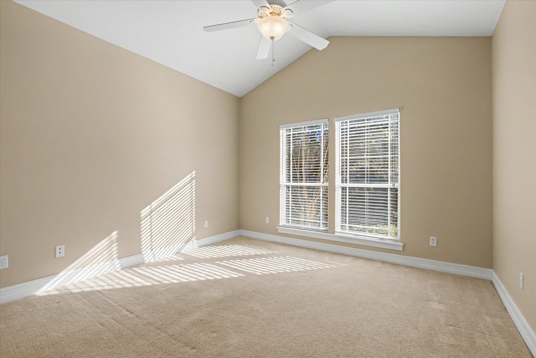 Empty bedroom with vaulted ceiling, ceiling fan, beige carpet, and two sunlit windows.