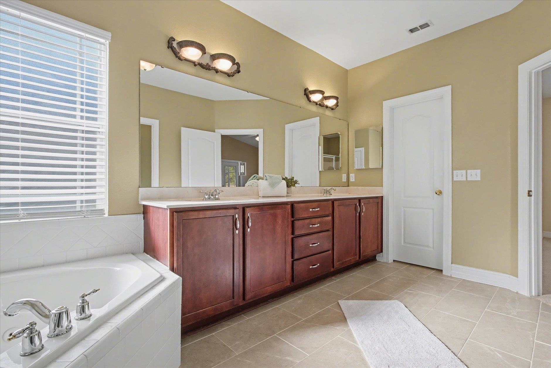 Bathroom with a long dark wood vanity, large mirror, and built-in soaking tub beneath a window.