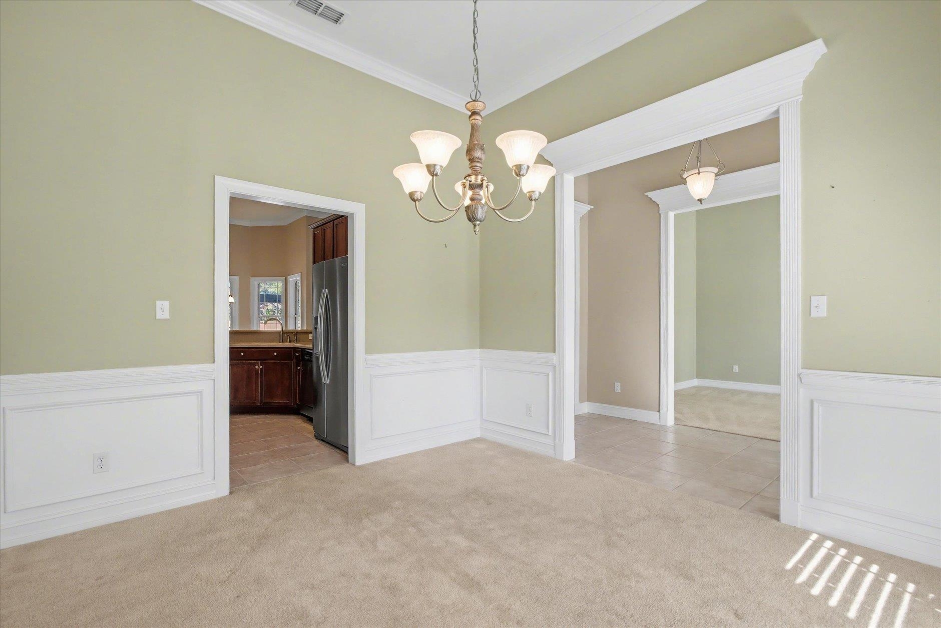 Empty dining room with paneled wainscoting, a chandelier, and openings to the kitchen and adjoining room.