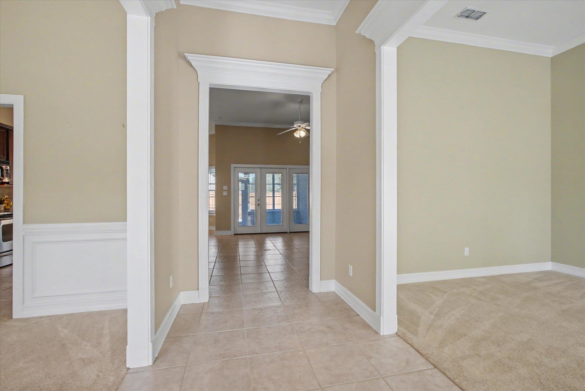 Tiled hallway with ornate white trim leading to glass-paneled French doors beside an empty carpeted room.