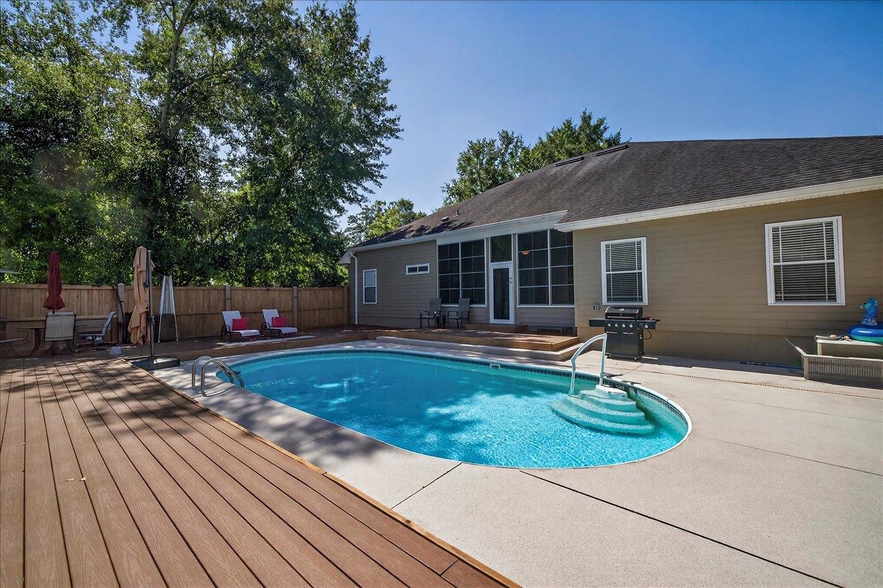 Backyard pool beside a tan house, with a broad wooden deck, fenced yard, and patio seating.
