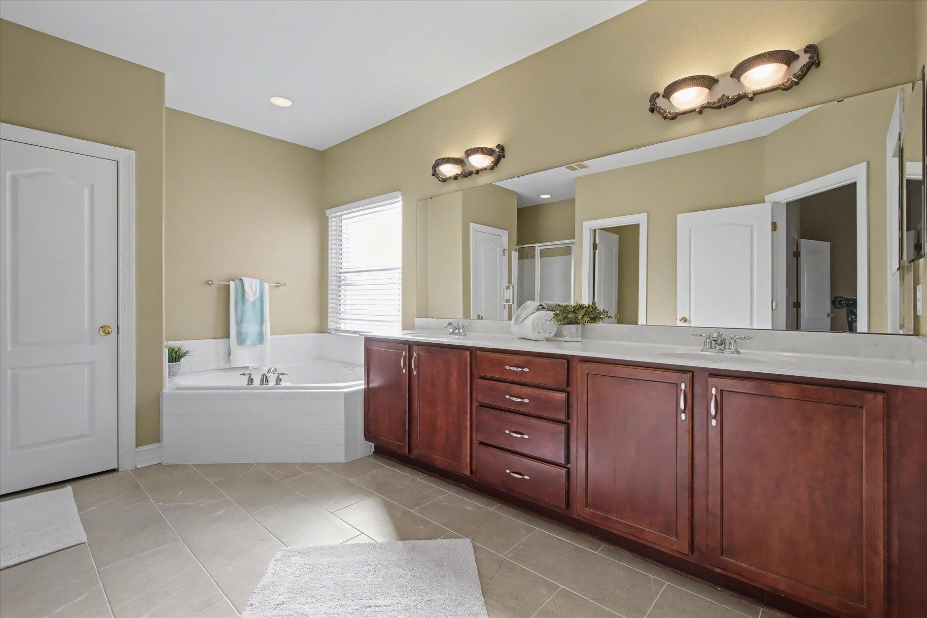 Bathroom with a long double vanity and a corner soaking tub beside a window.