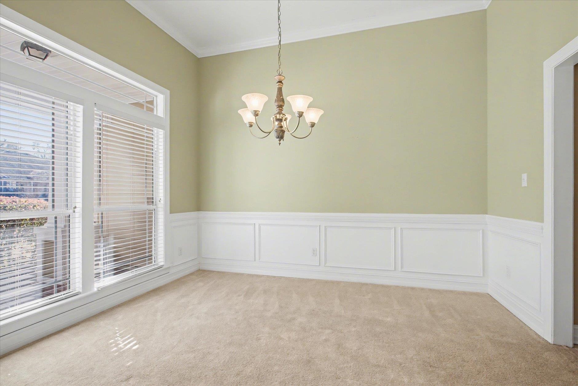 Empty dining room with white wainscoting, beige carpet, large windows, and a chandelier.