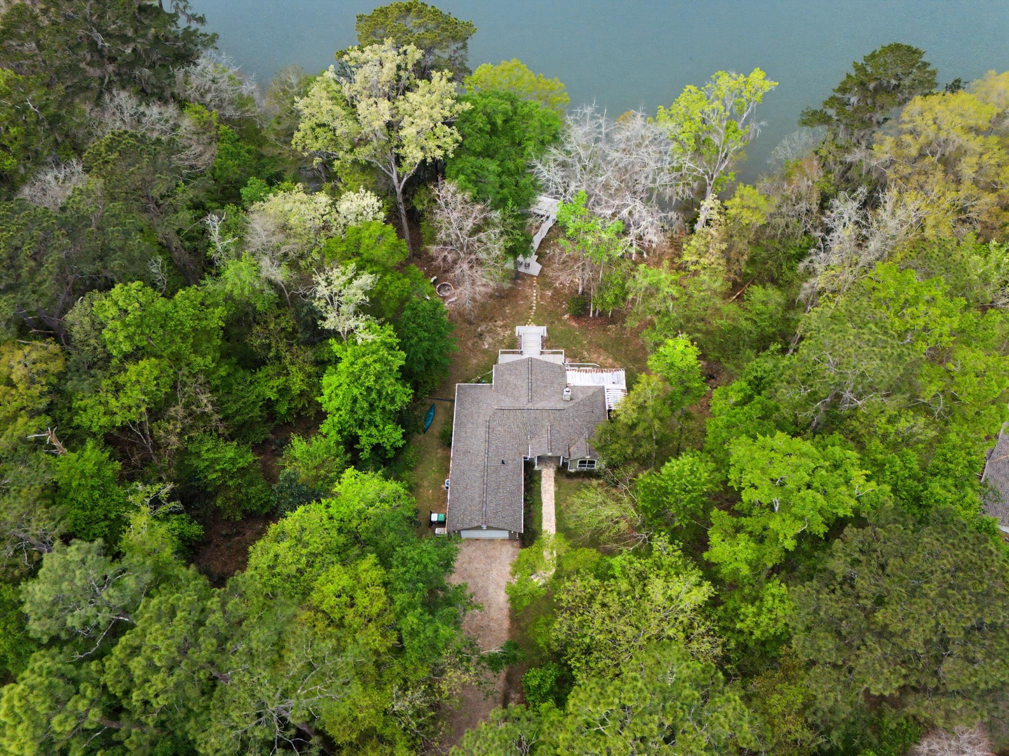 Aerial view of a wooded home beside a lake, with a dock and tree-lined yard.