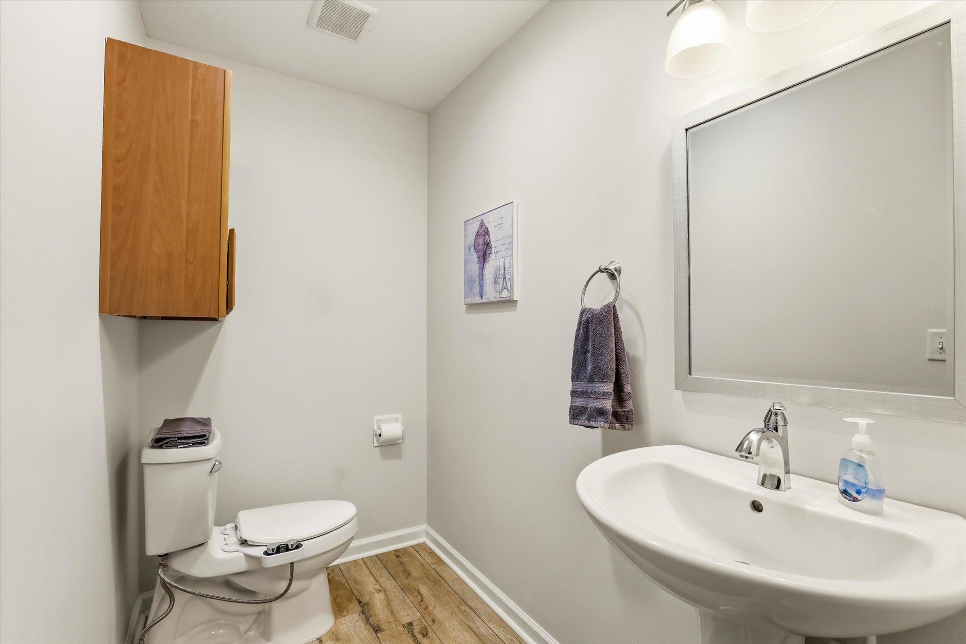 Bathroom with a pedestal sink, large mirror, toilet, and warm wood-look flooring.