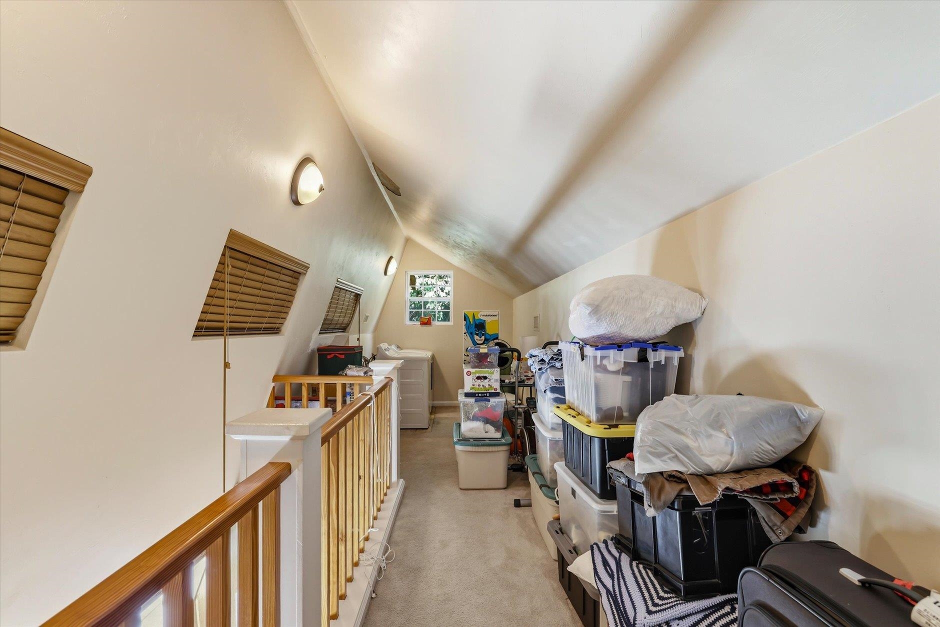 Carpeted upstairs storage loft with a wood railing, sloped ceiling, and stacked storage bins.