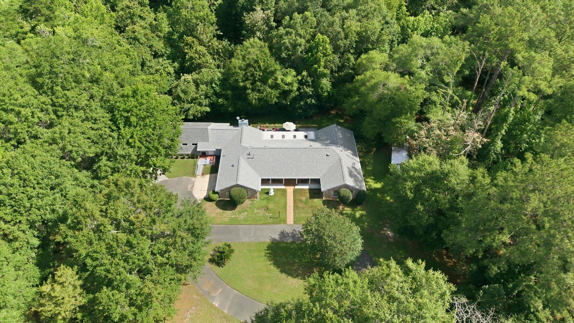 Aerial view of a single-story home surrounded by dense trees, with a curved driveway and front lawn.