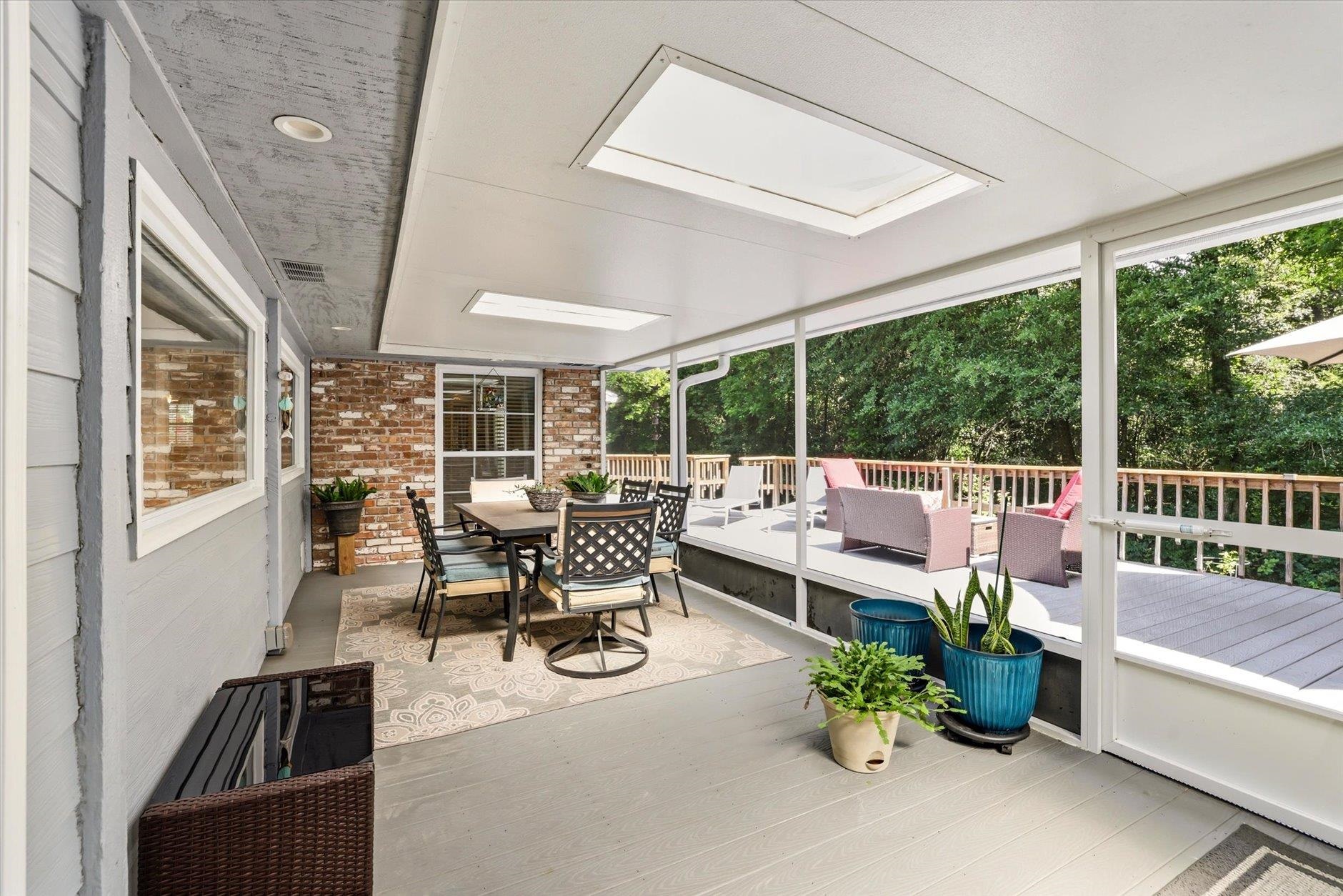 Screened porch with a dining set, skylights, potted plants, and leafy views of the deck.