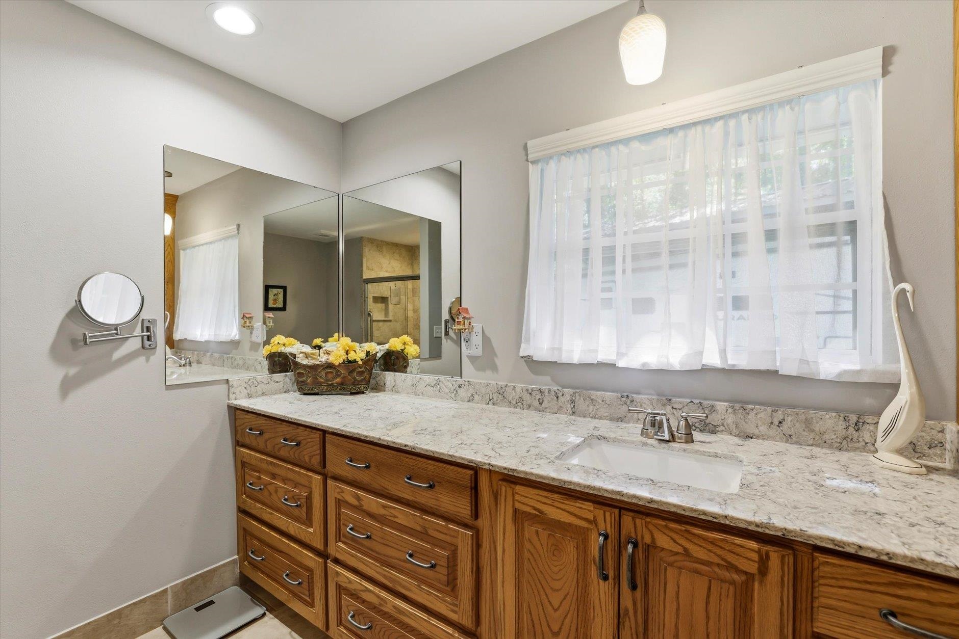 Bathroom vanity with a patterned stone countertop, wood cabinets, large mirrors, and a curtained window.