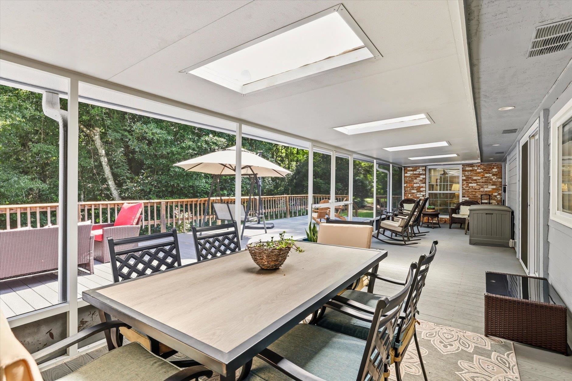 Screened porch with a long dining table, lounge seating, skylights, and leafy views.