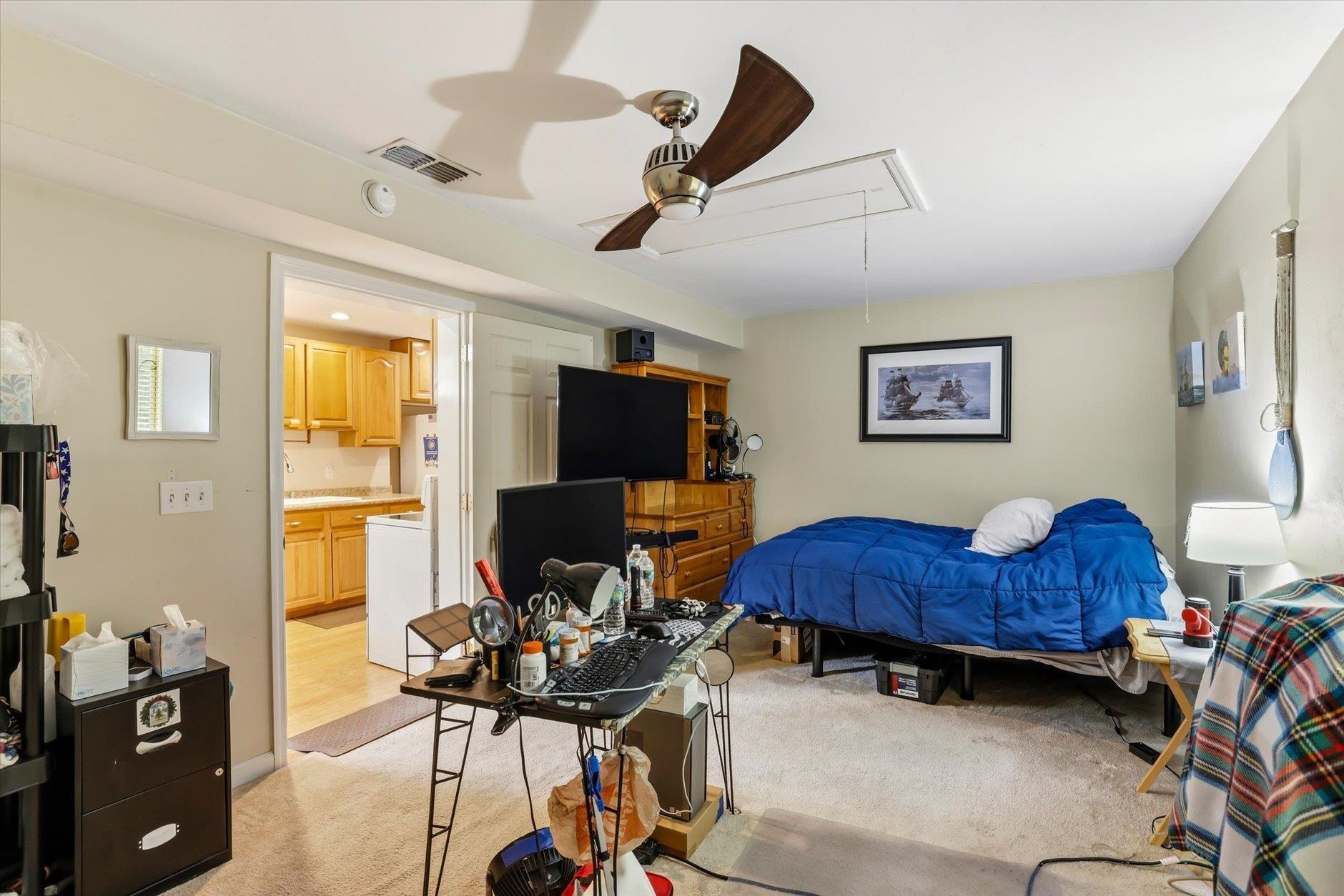 Bedroom with a blue-covered bed, ceiling fan, and desk; an open doorway leads to a compact kitchen.