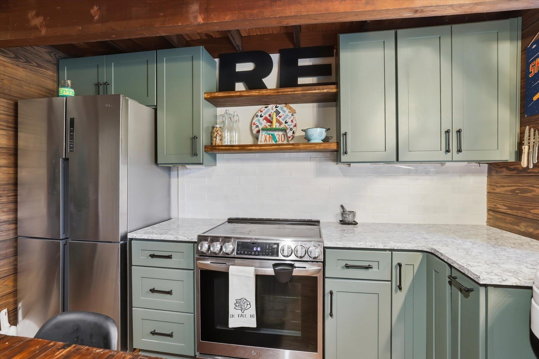 Kitchen with sage-green cabinets, wooden shelves, white tile backsplash, and stainless-steel appliances.